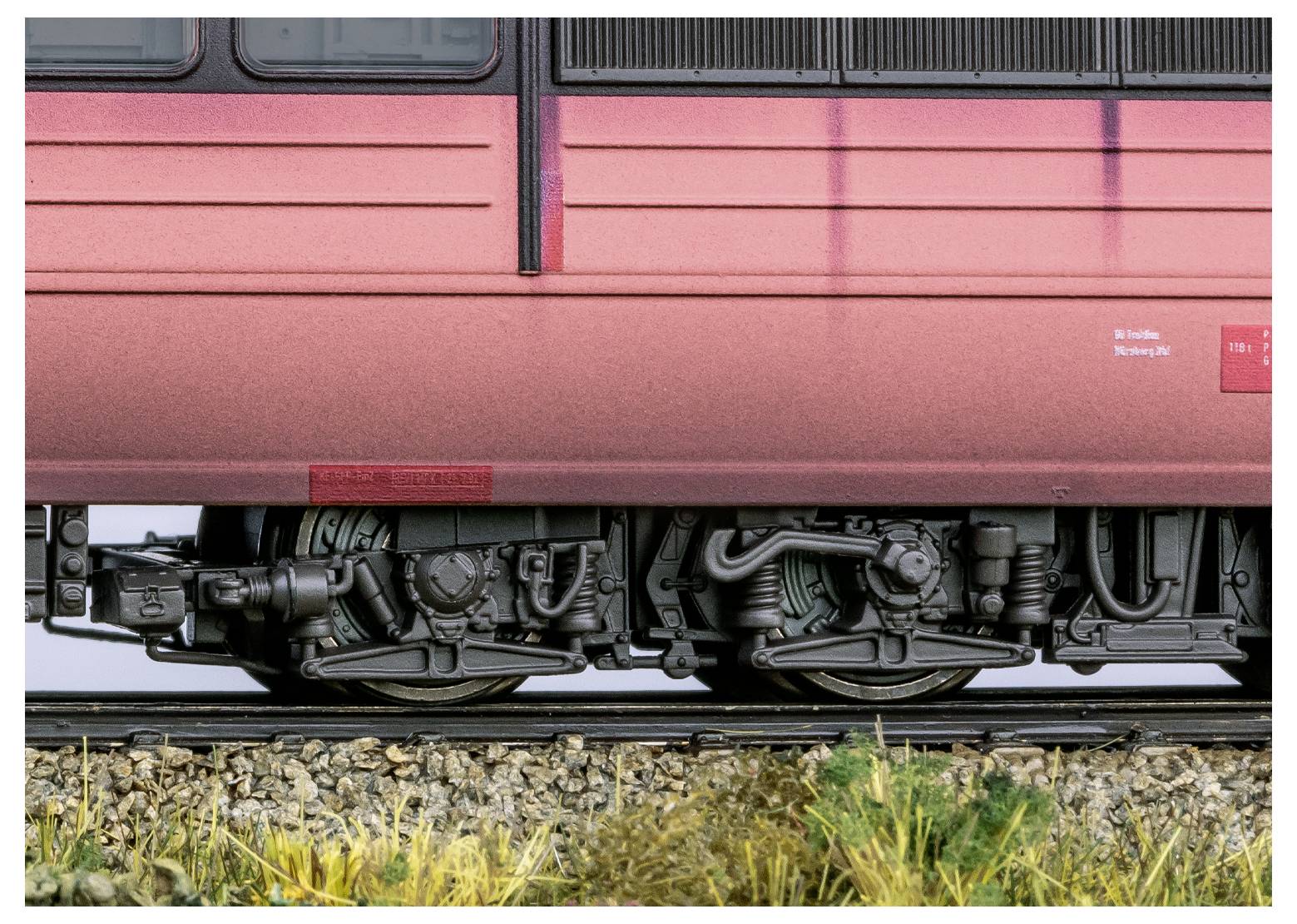 Detailaufnahme eines Zugrades auf Schienen, die feine Details des Untergestells vor einem unscharfen roten Zugäußeren zeigt.