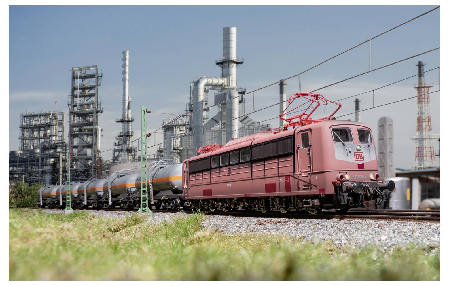 Ein roter Zug fährt auf Gleisen neben einem Industriewerk mit hohen Schornsteinen im Hintergrund, unter einem klaren blauen Himmel.