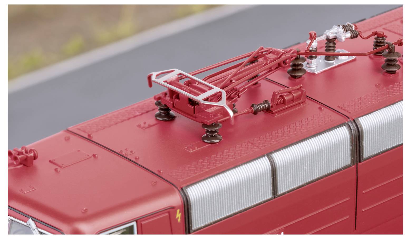 Draufsicht eines roten Modellzugdachs mit detaillierten elektrischen Komponenten und einem Stromabnehmer vor unscharfem Außenhintergrund.