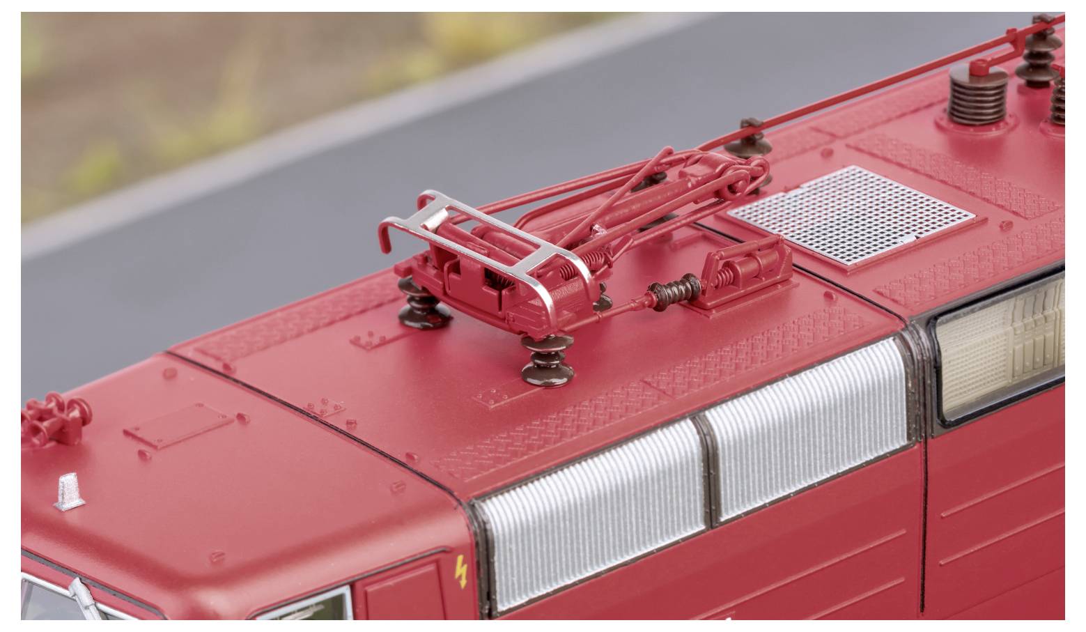 Das Bild zeigt das Dach einer roten elektrischen Lokomotive mit sichtbarem Stromabnehmer und Verkabelung vor einem unscharfen Hintergrund.