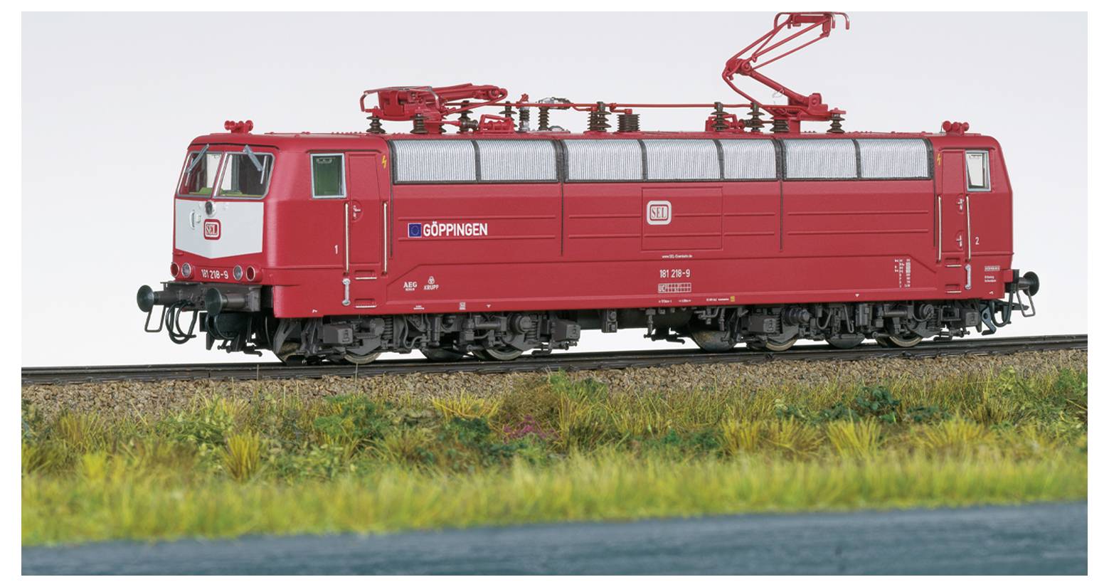Eine rote Lokomotive mit Oberleitung fährt auf einem Gleis, umgeben von grasiger Landschaft. Die Lokomotive trägt ein Schild mit der Aufschrift 