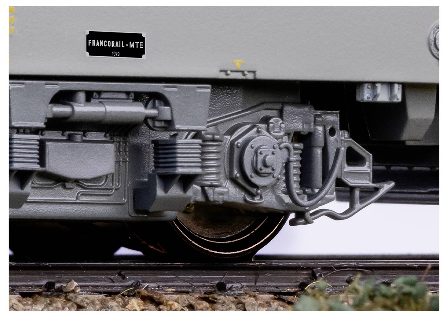 Nahaufnahme eines Modelleisenbahnrades und Federungsdetails auf einer Schiene, mit einem kleinen Schild im Hintergrund, auf dem 