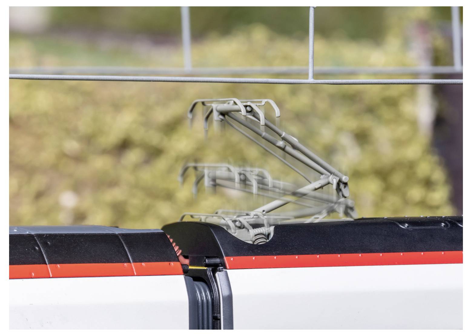 Ein Hochgeschwindigkeitszug-Dach mit einem verschwommenen Stromabnehmer, der Bewegung vor einem verschwommenen Hintergrund von Grünpflanzen andeutet.