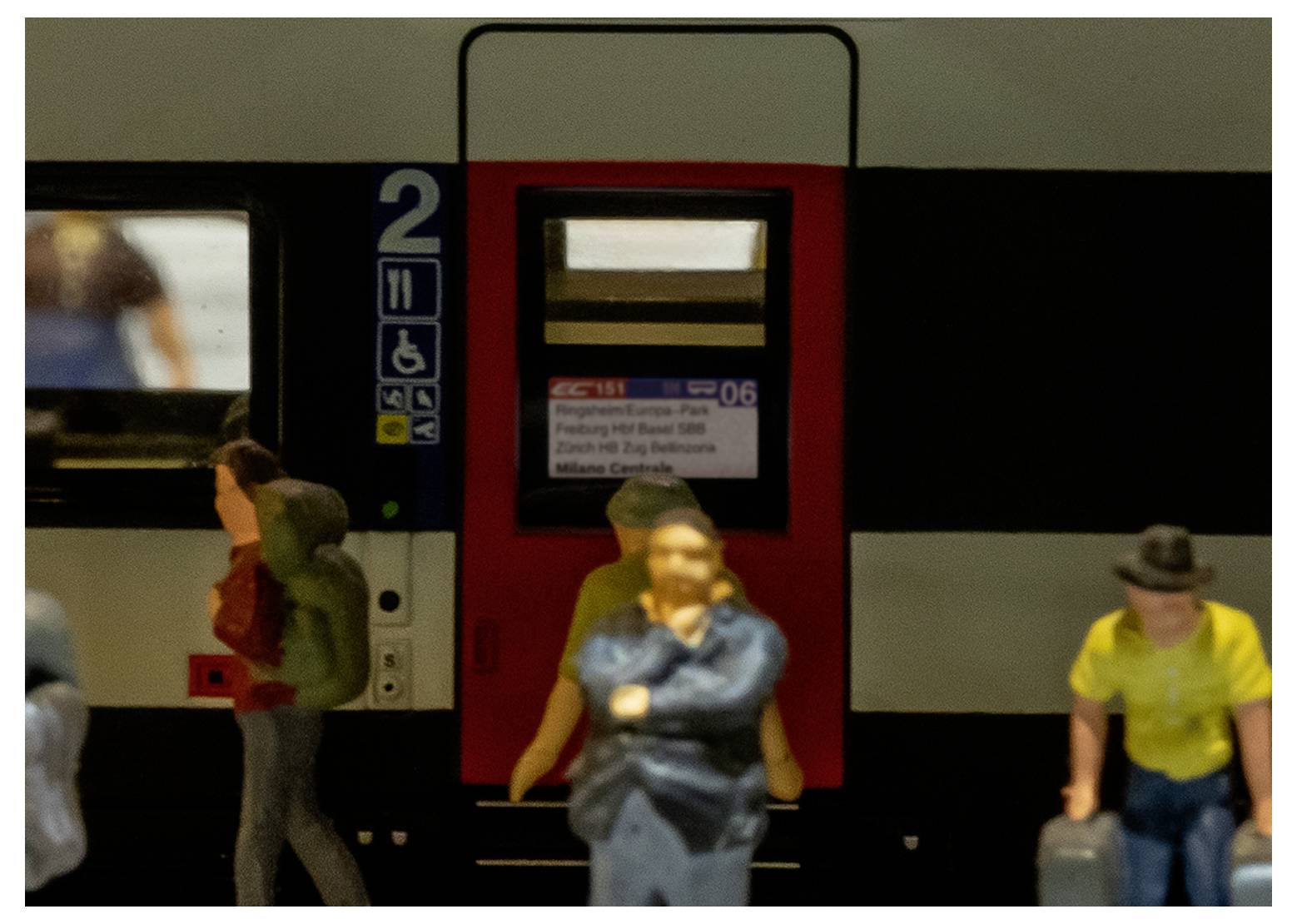 Ein Modellzug mit Figuren steht vor der Tür eines Eisenbahnwaggons. Die Waggonnummer '2' und verschiedene Symbole weisen auf Barrierefreiheitsfunktionen hin.