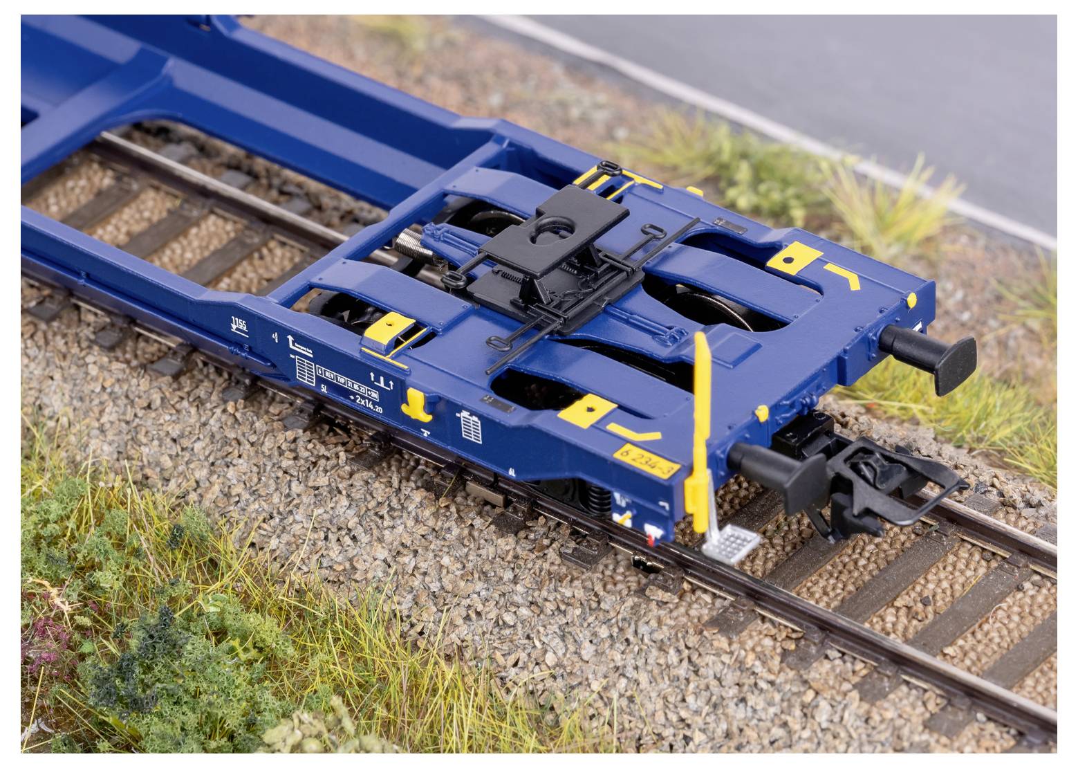 Ein detailliertes Skalenmodell eines blauen Eisenbahnwagenrahmens auf einer Schiene, das Kupplungsmechanismen sowie umgebendes Kies und Grünpflanzen zeigt.