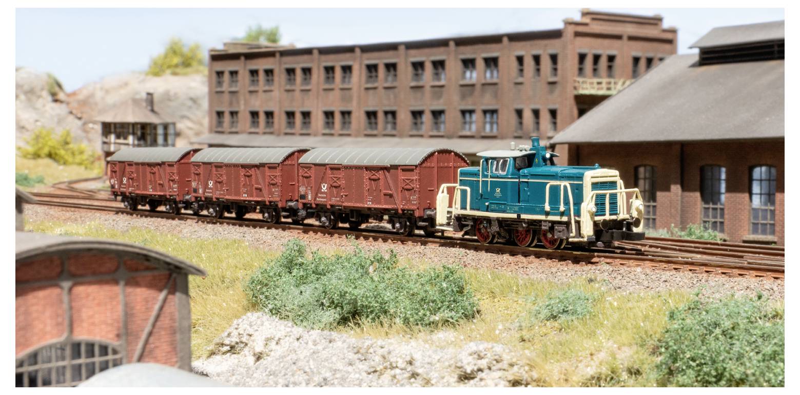 Ein Modelleisenbagzug mit einer blauen Lokomotive und roten Güterwaggons fährt an einem Backsteingebäude in Industrieoptik und grüner Landschaft in einer Miniaturlandschaft vorbei.