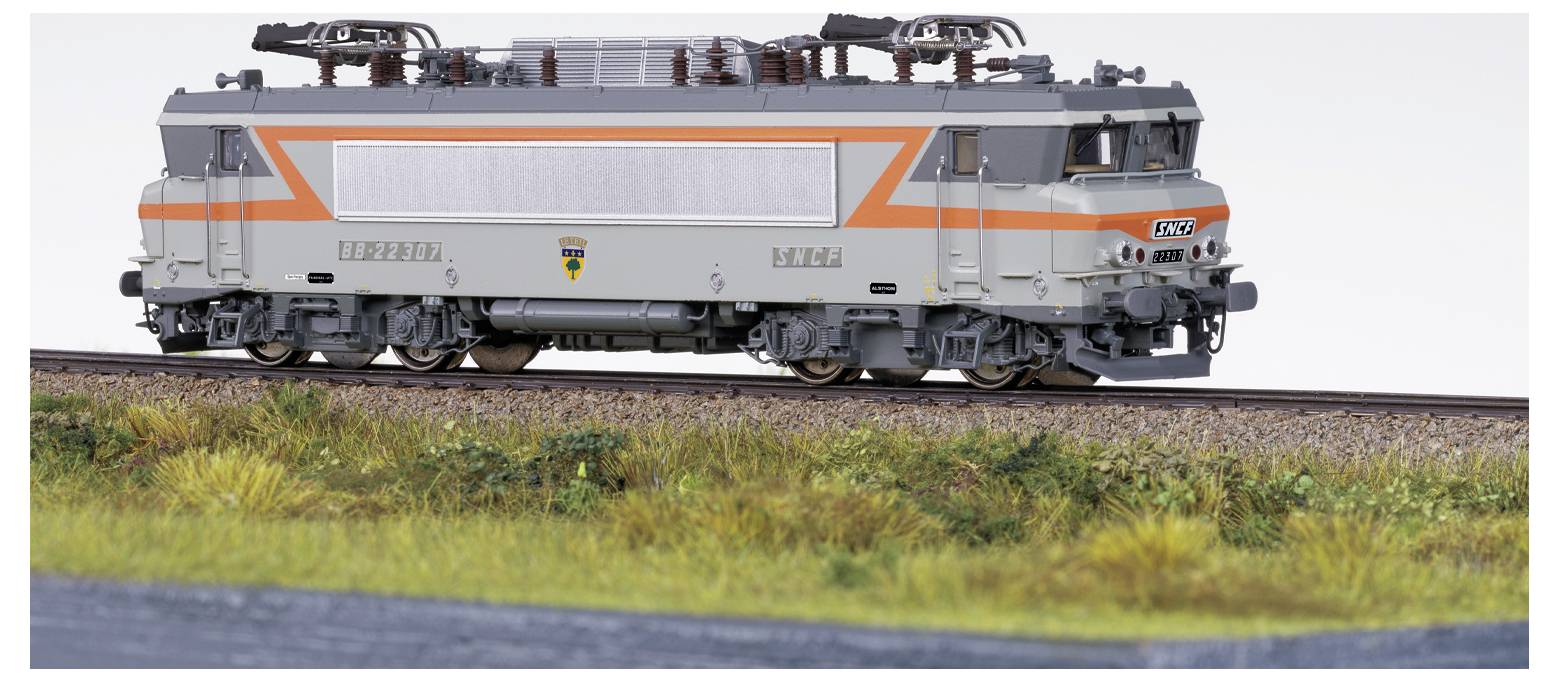 Eine Modelleisenbok mit grau-oranger Farbgebung, ausgestellt und auf einer Schiene fahrend, umgeben von Gras und Landschaft.