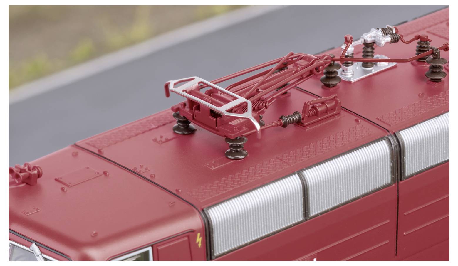 Eine detaillierte Ansicht des Daches einer roten elektrischen Lokomotive, die sich auf den Stromabnehmer und die Isolatoren vor einem unscharfen Gleishintergrund konzentriert.