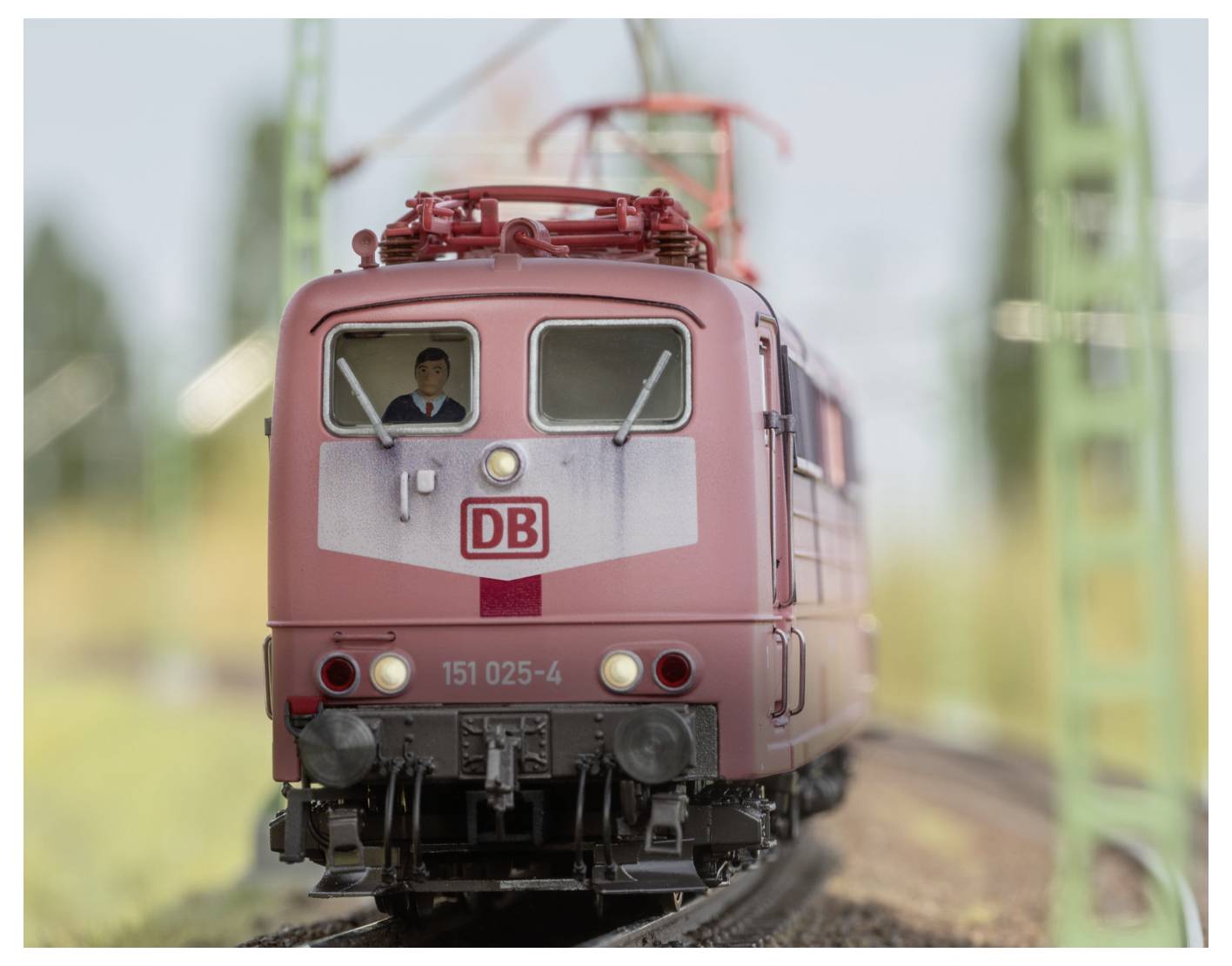 Ein Modelleisenbahnzug mit 'DB'-Logo auf der Front bewegt sich auf einer Schiene, gefahren von einem sichtbaren Führer. Grüne Strukturen und verschwommene Landschaft im Hintergrund.
