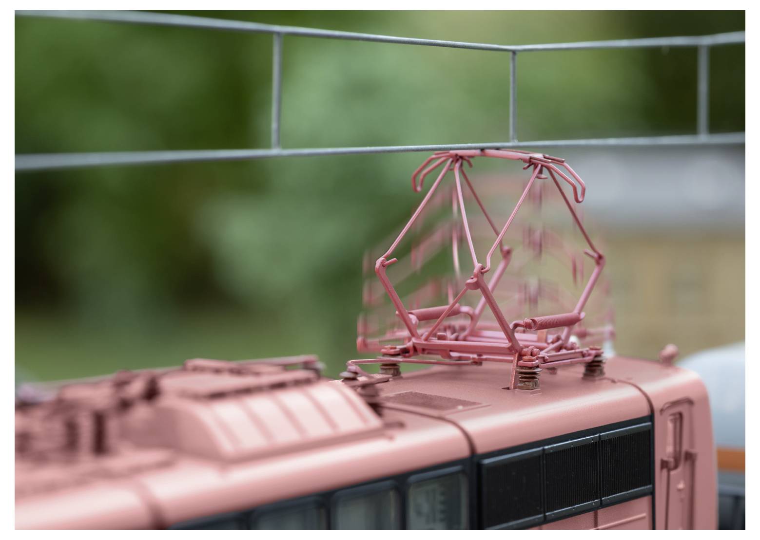 Nahaufnahme des Daches eines Modelleisenbahnwagens mit komplexer roter Elektroausrüstung vor einem unscharfen grünen Hintergrund.