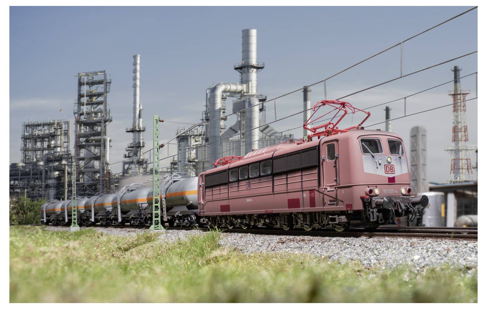 Ein Modellzug mit einer roten Lokomotive und Kesselwagen fährt auf Schienen vor einer detaillierten industriellen Raffinerielandschaft unter einem klaren Himmel.