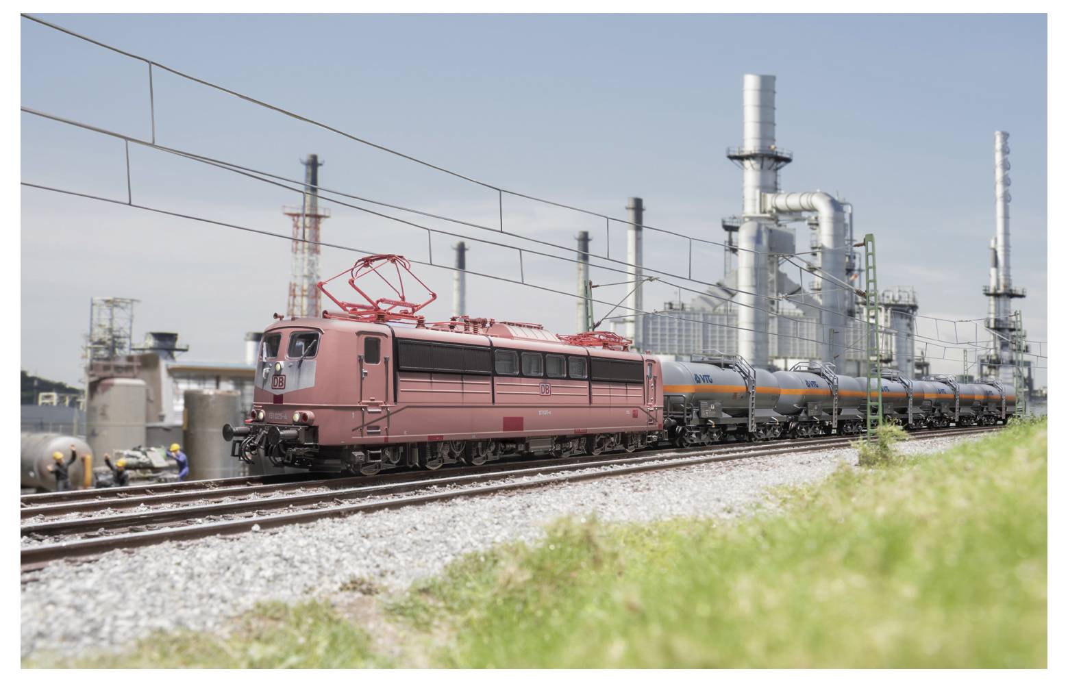 Ein Zug mit roten Lokomotiven und industriellen Tankwagen fährt entlang der Gleise durch ein Industriegebiet mit großen Schornsteinen im Hintergrund.