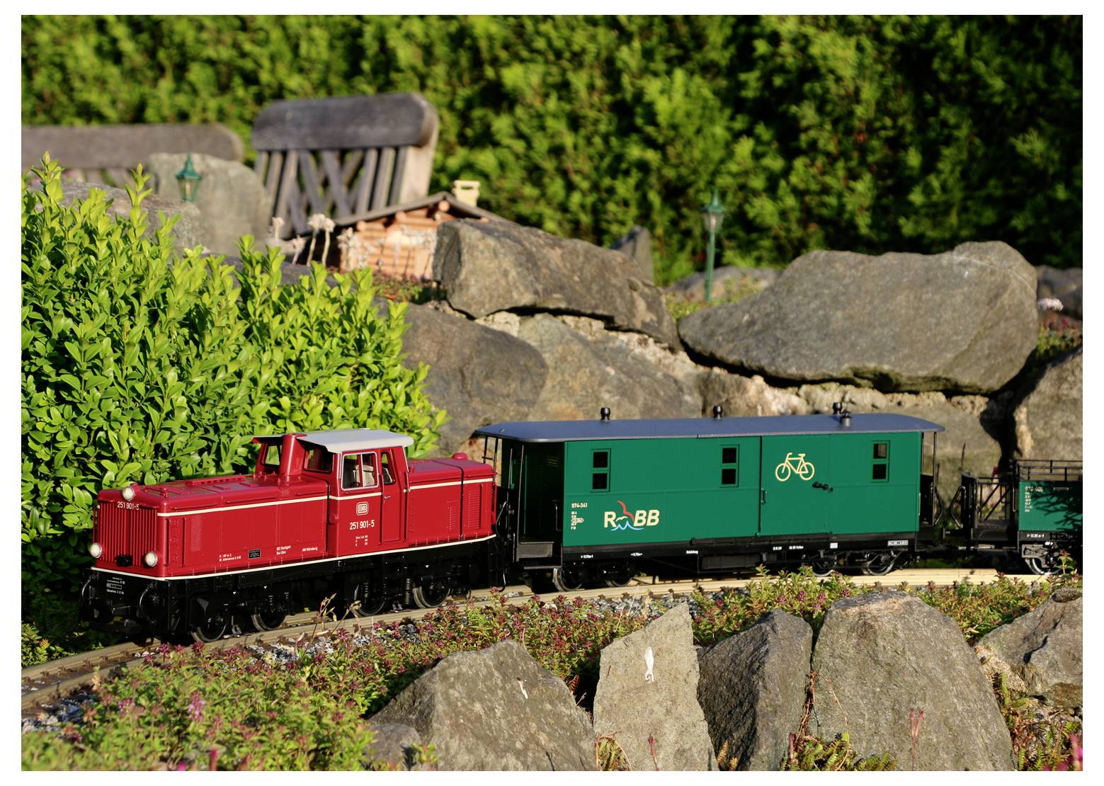 Eine Modell-Lokomotive in Rot zieht einen grünen Güterwaggon mit einem Fahrrad-Symbol, der durch eine Gartenszene mit Steinen und Grünpflanzen fährt.