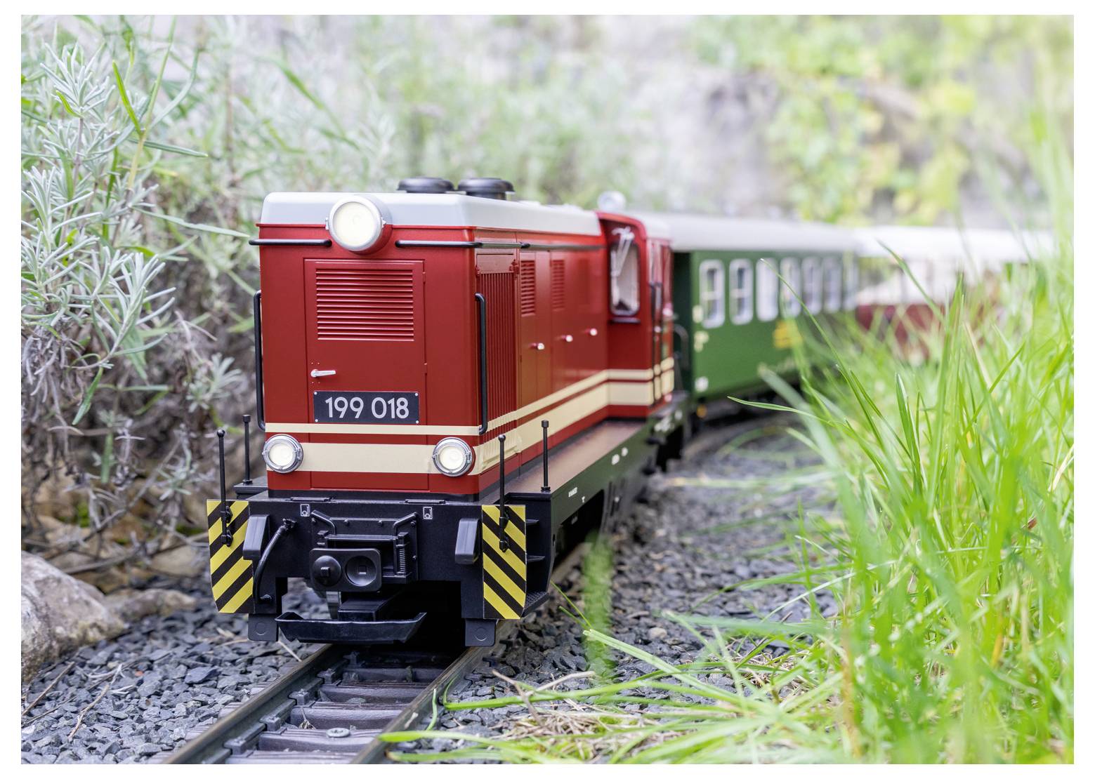 Ein roter Modelleisenbahn mit der Aufschrift '199 018' auf der Front fährt auf einer Strecke, die von Grünpflanzen umgeben ist.