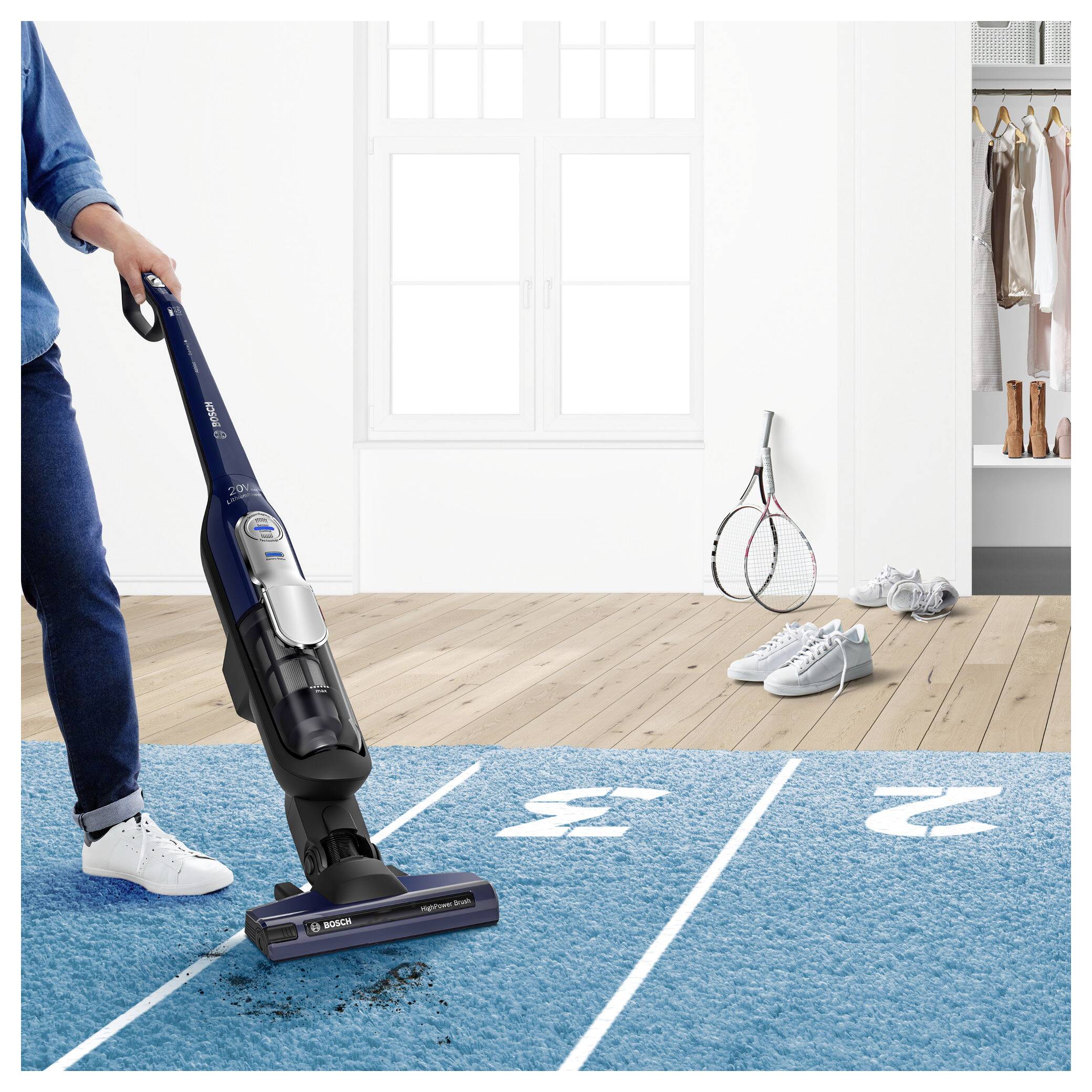 Eine Person saugt einen Teppich ab, der als 100-Meter-Rennstrecke bemalt ist, während Tennisausrüstung auf dem Holzboden daneben liegt.