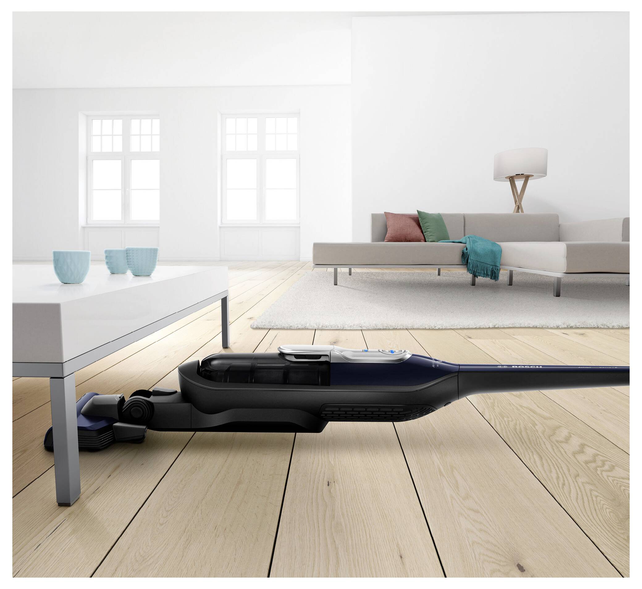 Ein Staubsauger liegt flach, um unter einem weißen Couchtisch in einem hellen Wohnzimmer zu reinigen, während eine Couch und eine Lampe im Hintergrund zu sehen sind.