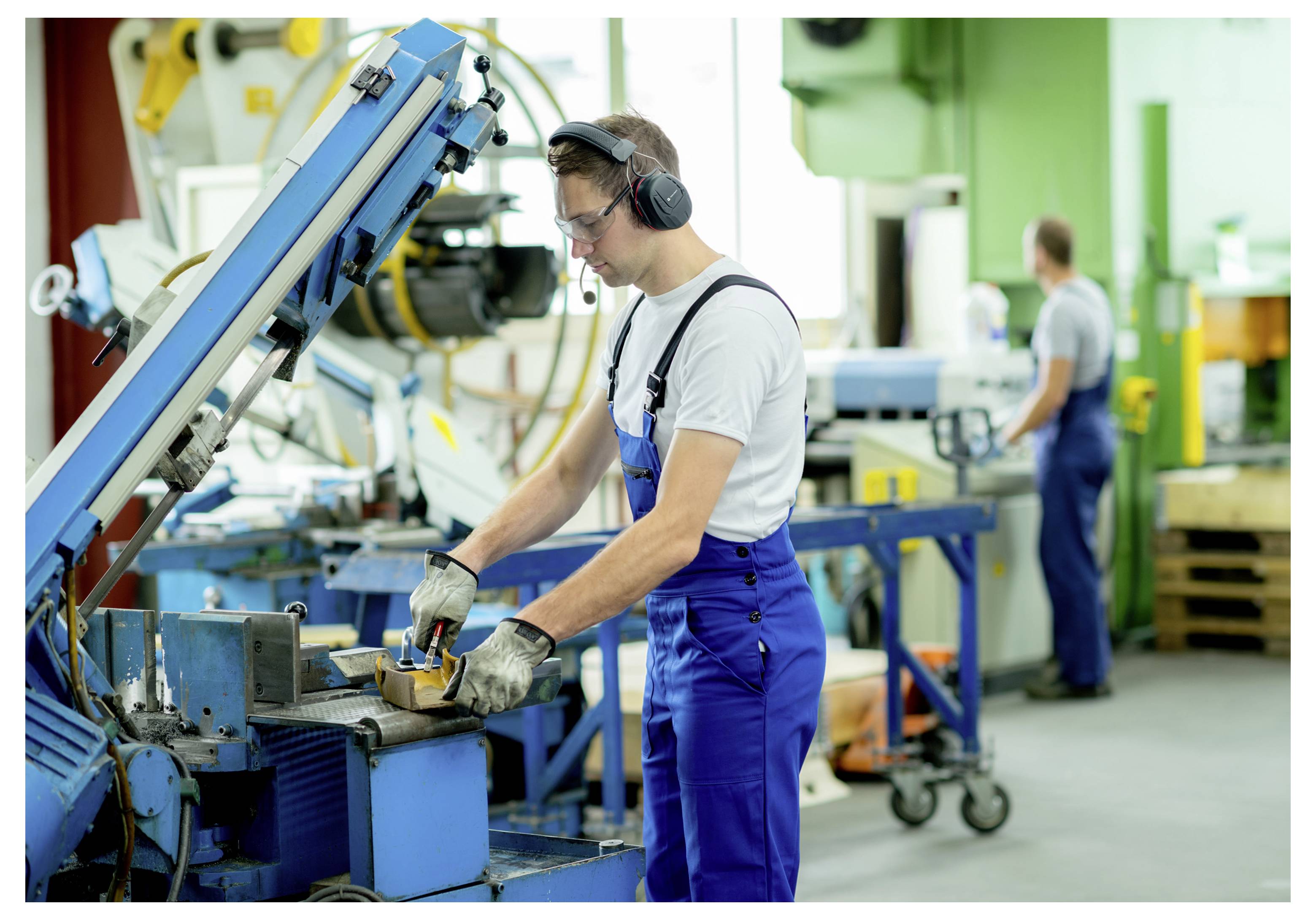 Fabrikarbeiter in blauer Arbeitskleidung und Kopfhörern bedient eine blaue Industriemaschine in einer Werkstatt, während ein weiterer Arbeiter im Hintergrund verschwommen zu sehen ist.