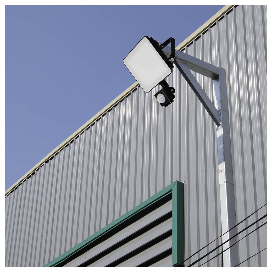 Eine Außenflutlampe, die an einem gewellten Metallgebäude unter einem klaren blauen Himmel montiert ist und Sicherheit oder nächtliche Beleuchtung hervorhebt.