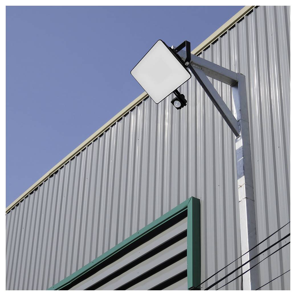 Eine moderne Industriegebäudefassade mit vertikaler Metallverkleidung, ausgestattet mit einer montierten quadratischen LED-Leuchte und einer Sicherheitskamera vor einem klaren Himmel.