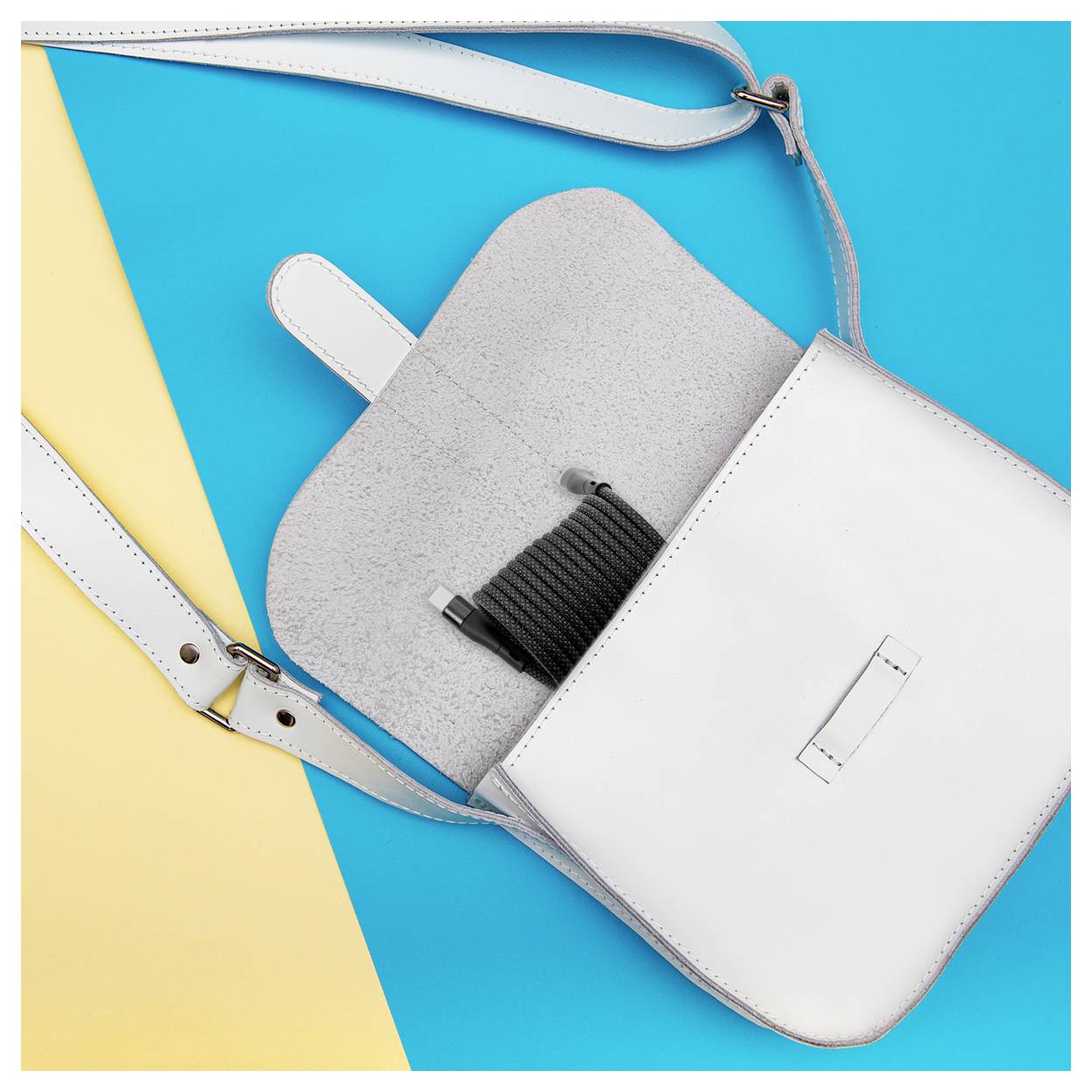 Eine weiße Lederhandtasche mit einem beigen Riemen, die vor einem blau-gelben Hintergrund geöffnet ist und eine aufgewickelte schwarze Schnur im Inneren zeigt.