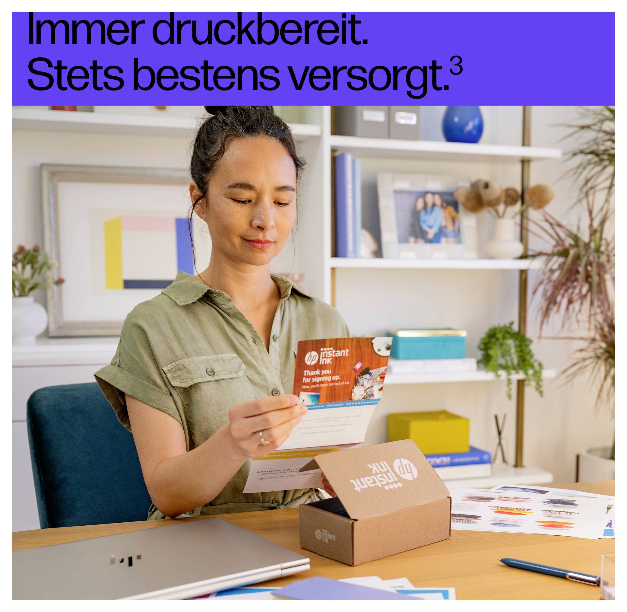 Eine Frau sitzt an einem Schreibtisch und packt HP Instant Ink Patronen aus. Ein Laptop und farbige Drucke sind zu sehen. Text oben liest: 'Immer druckbereit. Stets bestens versorgt.'