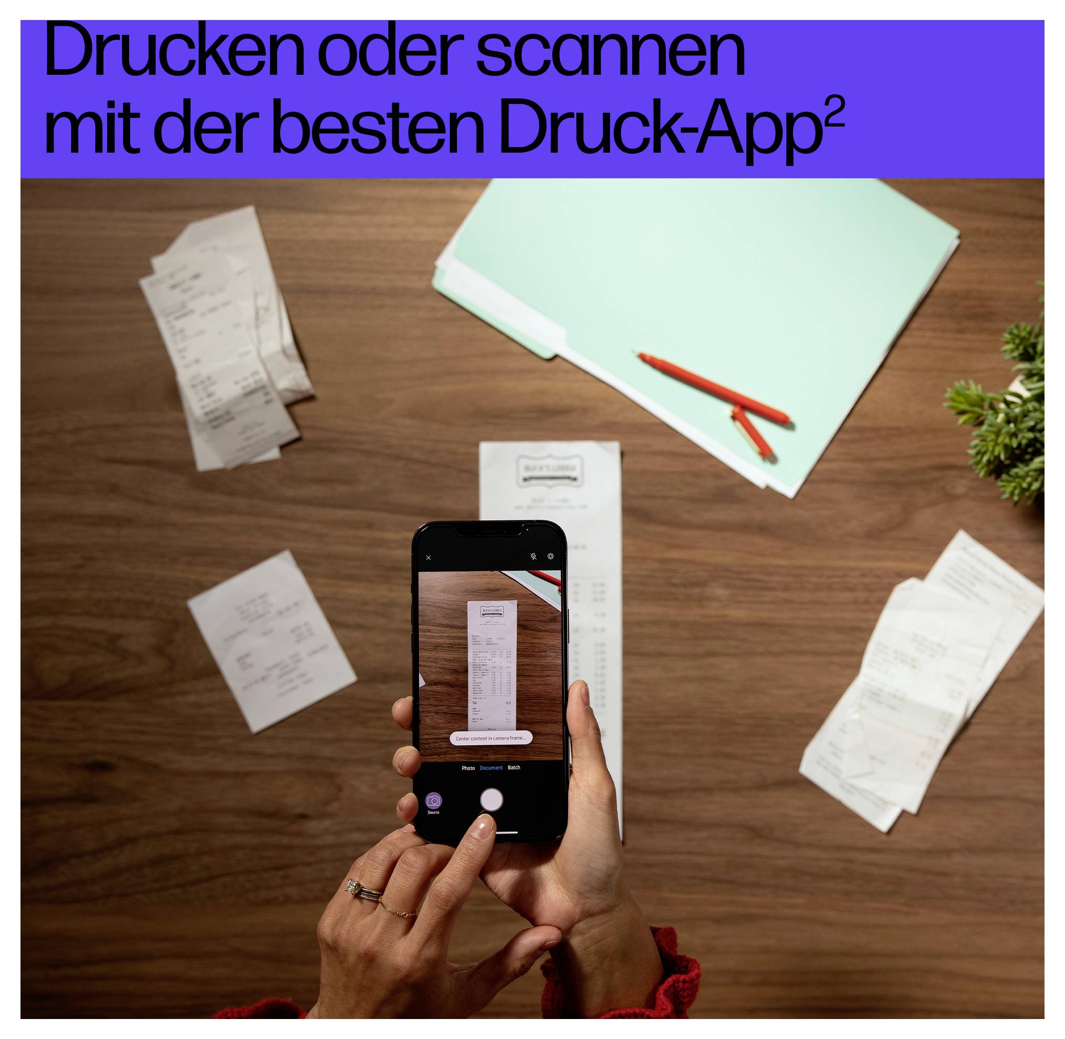 Person, die einen Kassenbon mit einem Smartphone auf einem Holzschreibtisch scannt, umgeben von Dokumenten und einem hellgrünen Ordner. Text auf Deutsch bewirbt eine Druck-App.