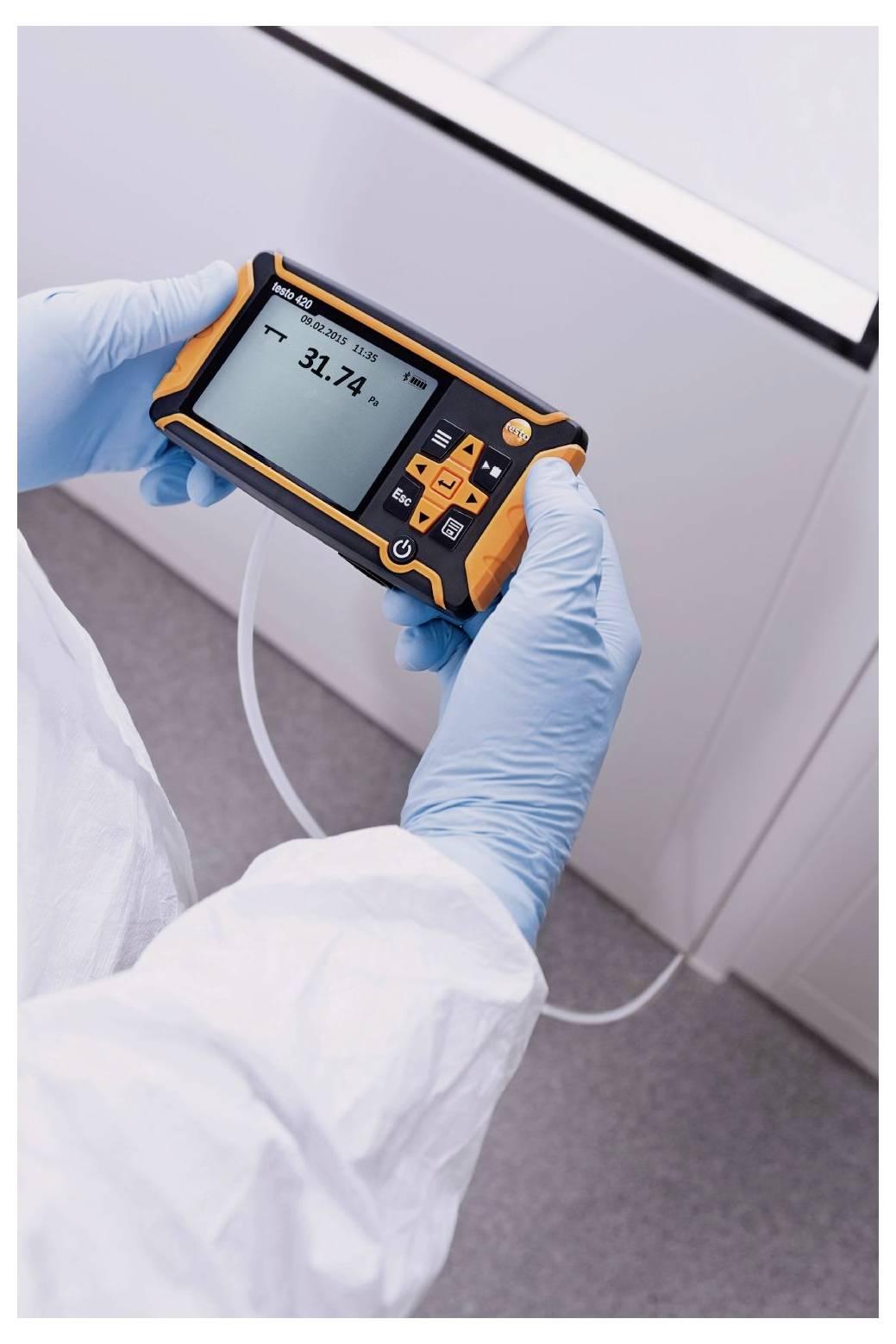 Eine Person in Schutzausrüstung hält ein digitales Gerät, das '31,2°C' anzeigt und möglicherweise Temperatur misst, verbunden mit Schläuchen in einer Laborumgebung.