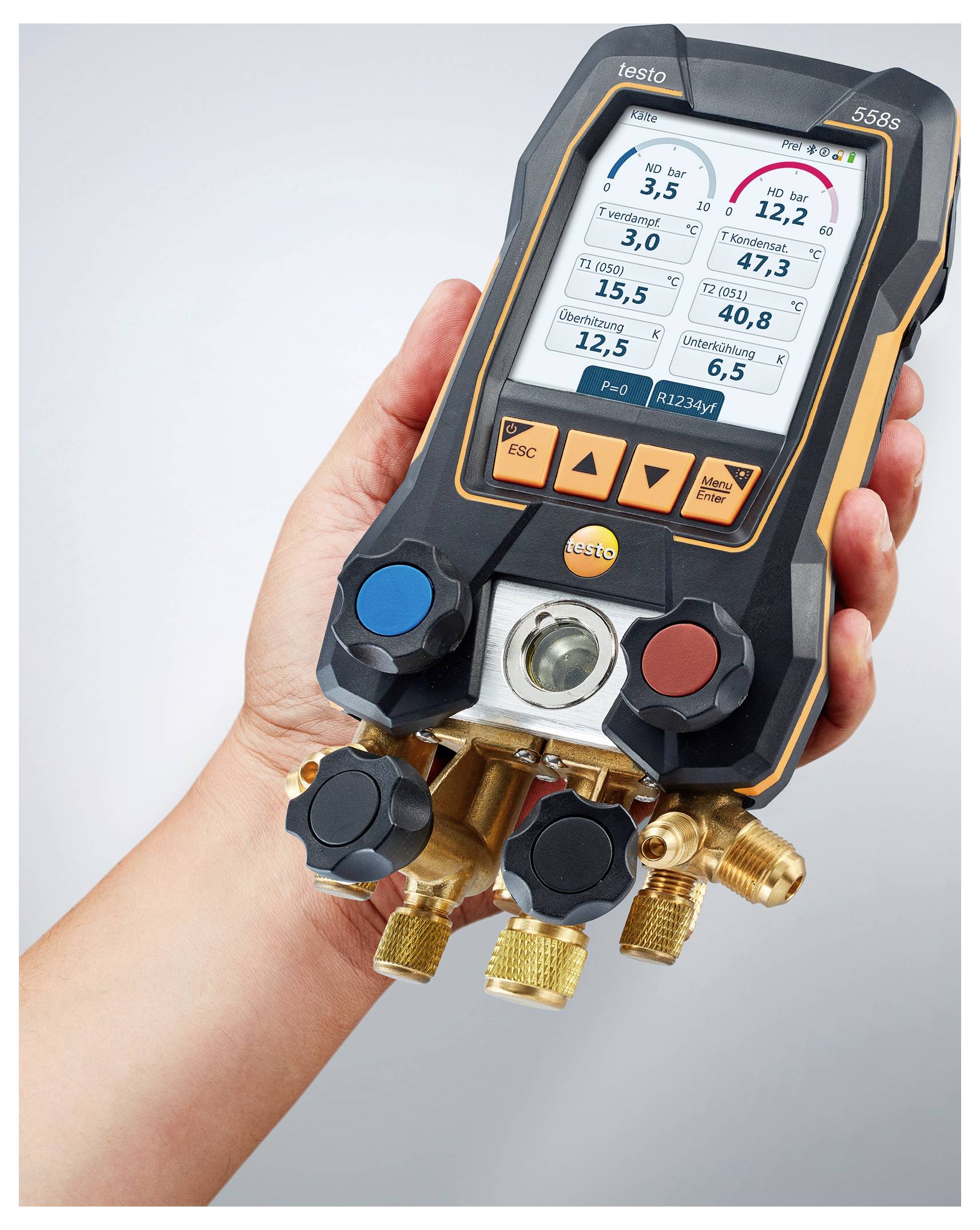Eine Hand, die einen digitalen Sammler mit Manometer hält, der Druck- und Temperaturanzeigen auf dem Bildschirm zeigt. Schwarzes und orangefarbenes Gerät mit mehreren Drehknöpfen.