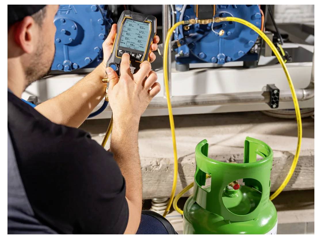 Eine Person verwendet ein digitales Gerät, um Messwerte zu überprüfen, höchstwahrscheinlich Temperatur oder Druck, mit einem grünen Gaszylinder in der Nähe in einer industriellen Umgebung.