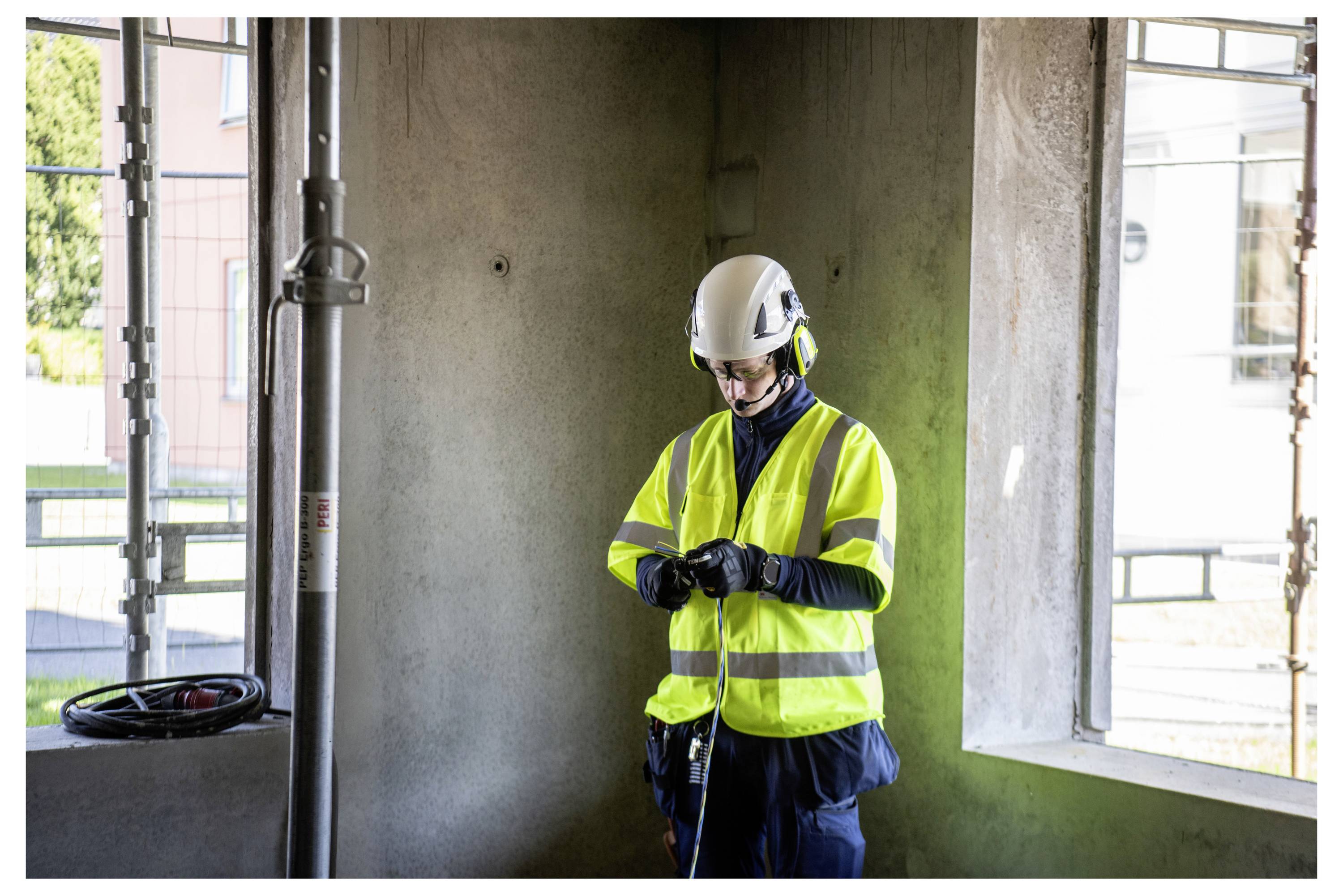 Ein Bauarbeiter in einer Sicherheitsweste und mit Helm steht drinnen in der Nähe von Fenstern und überprüft Ausrüstung.