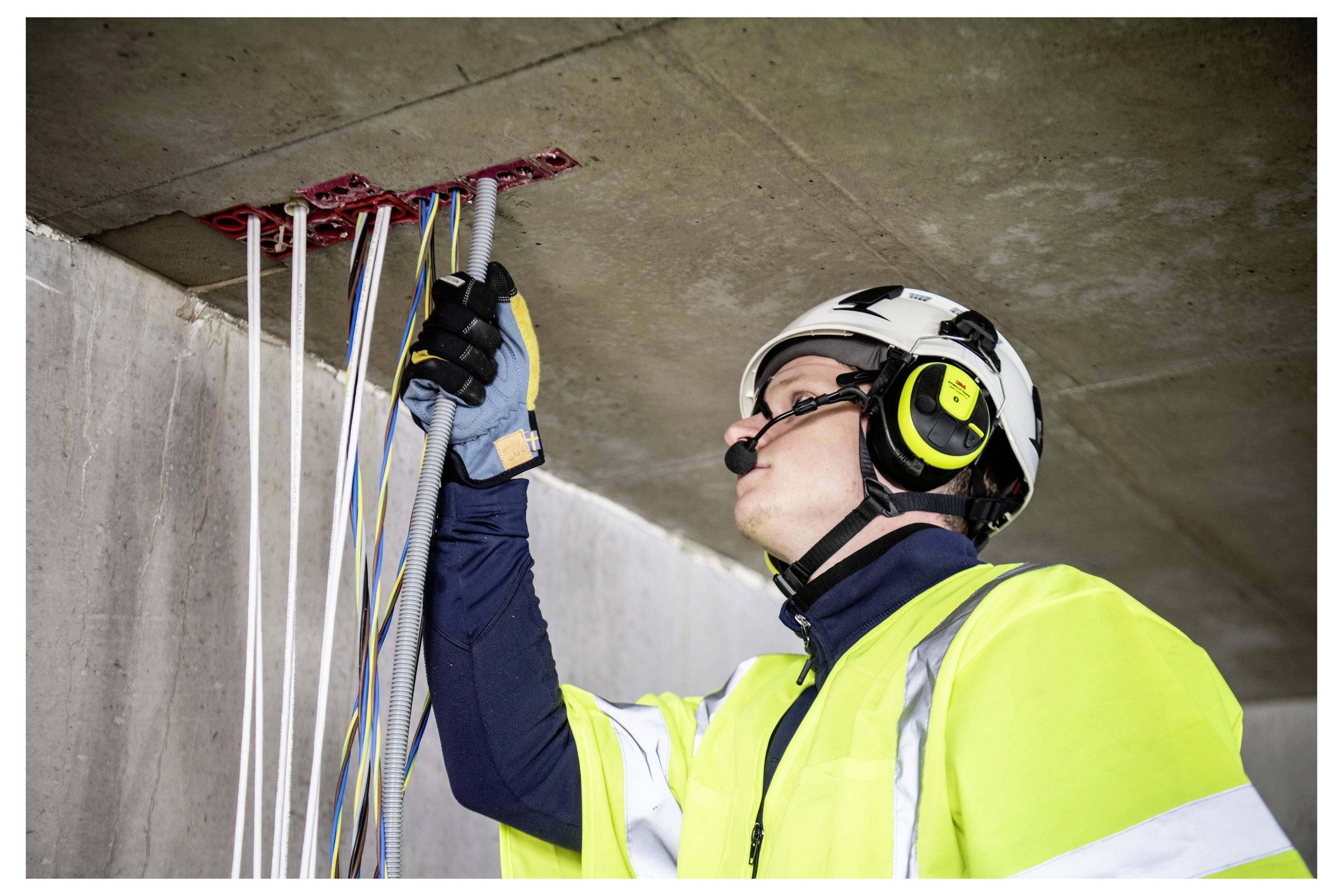 Ein Bauarbeiter in Warnschutzkleidung und Helm inspiziert Kabel, die aus einer Decke herausragen, und trägt dabei Gehörschutz und Schutzhandschuhe.