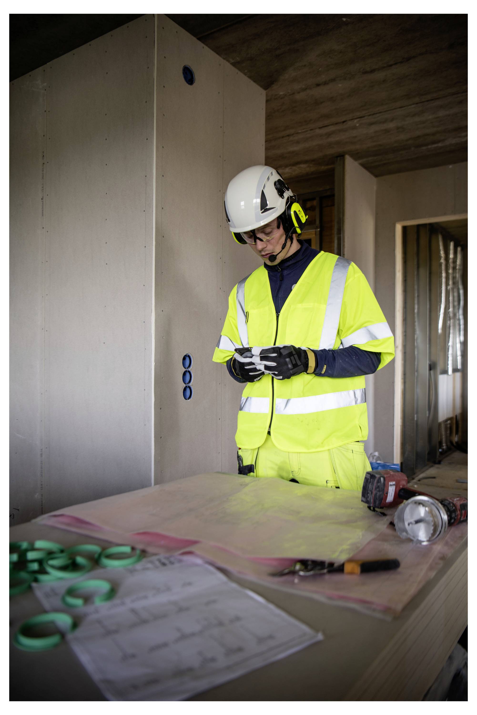 Ein Bauarbeiter in Warnschutzkleidung studiert ein Papier in einem Gebäude im Bau. Werkzeuge und Materialien liegen auf einem nahe gelegenen Tisch.