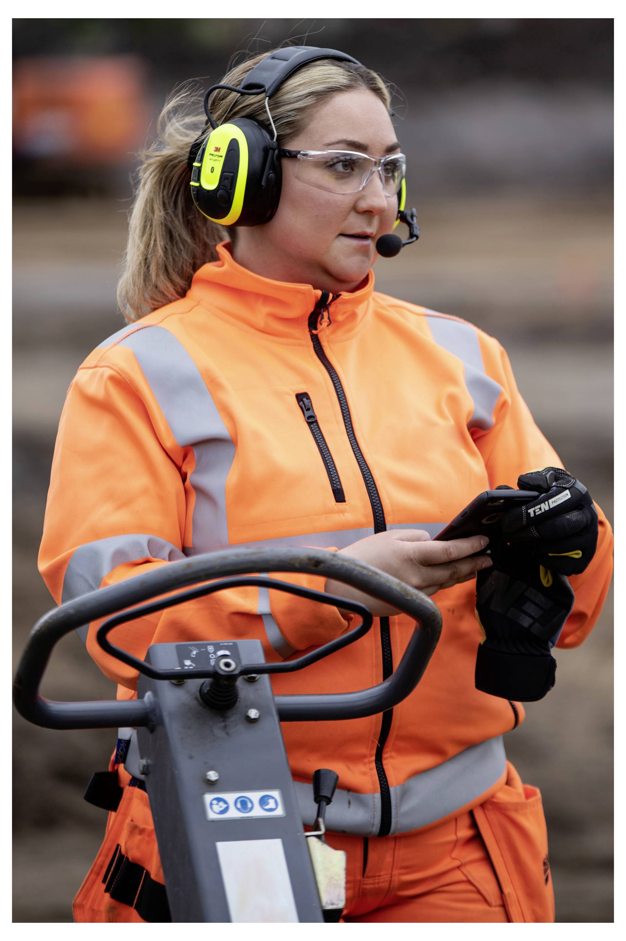 Eine Person, die eine orangefarbene Warnweste und Schutzausrüstung trägt, einschließlich Kopfhörern und Sicherheitsbrille, bedient eine Maschine auf einer Baustelle.