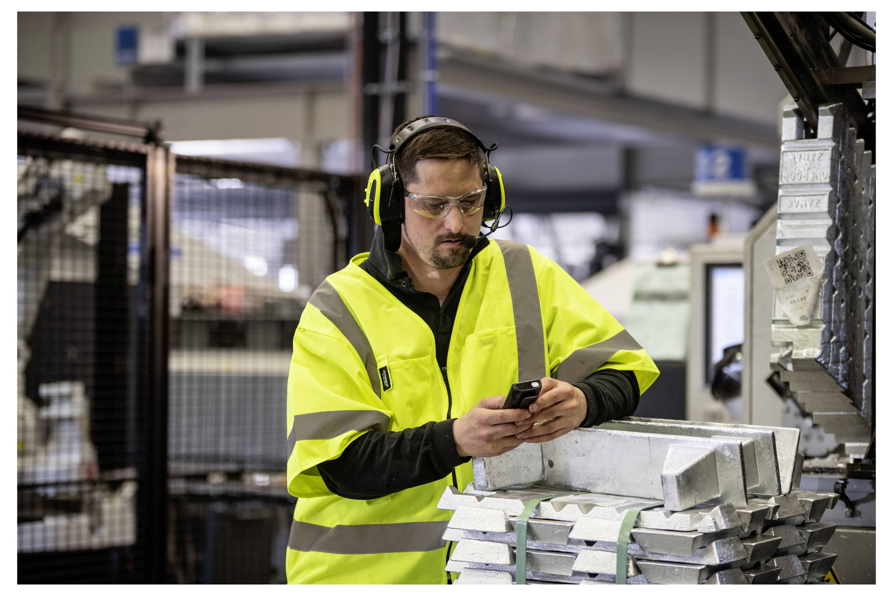 Ein Arbeiter in einer Warnweste und Sicherheitsausrüstung prüft sein Handy, während er neben Metallmaterialien in einer industriellen Umgebung steht.