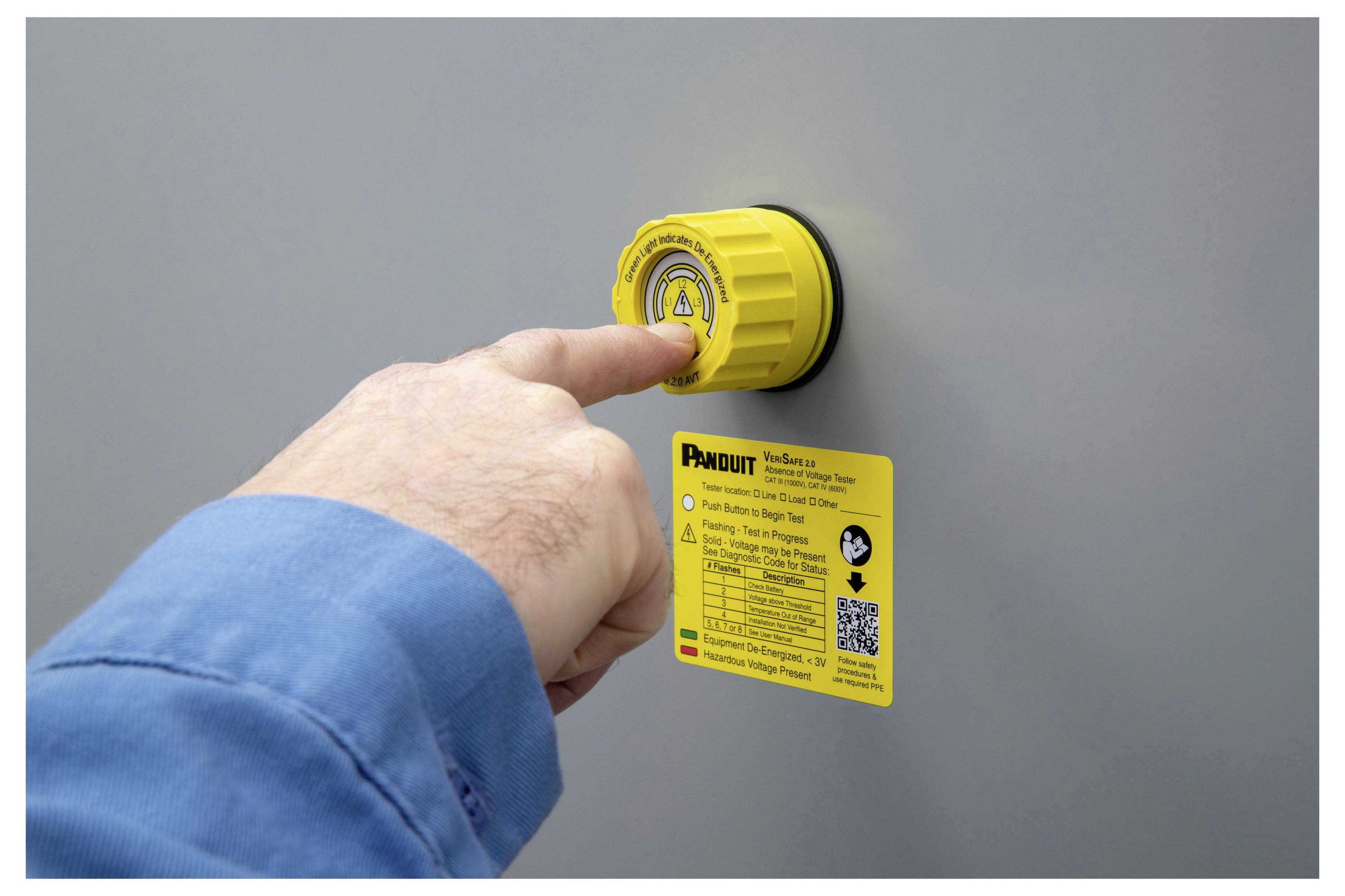 Eine Hand drückt ein gelbes Sperrgerät mit einem Sicherheitsetikett auf einer grauen Oberfläche. Das Etikett enthält Anweisungen und einen QR-Code für Sicherheitsinformationen.