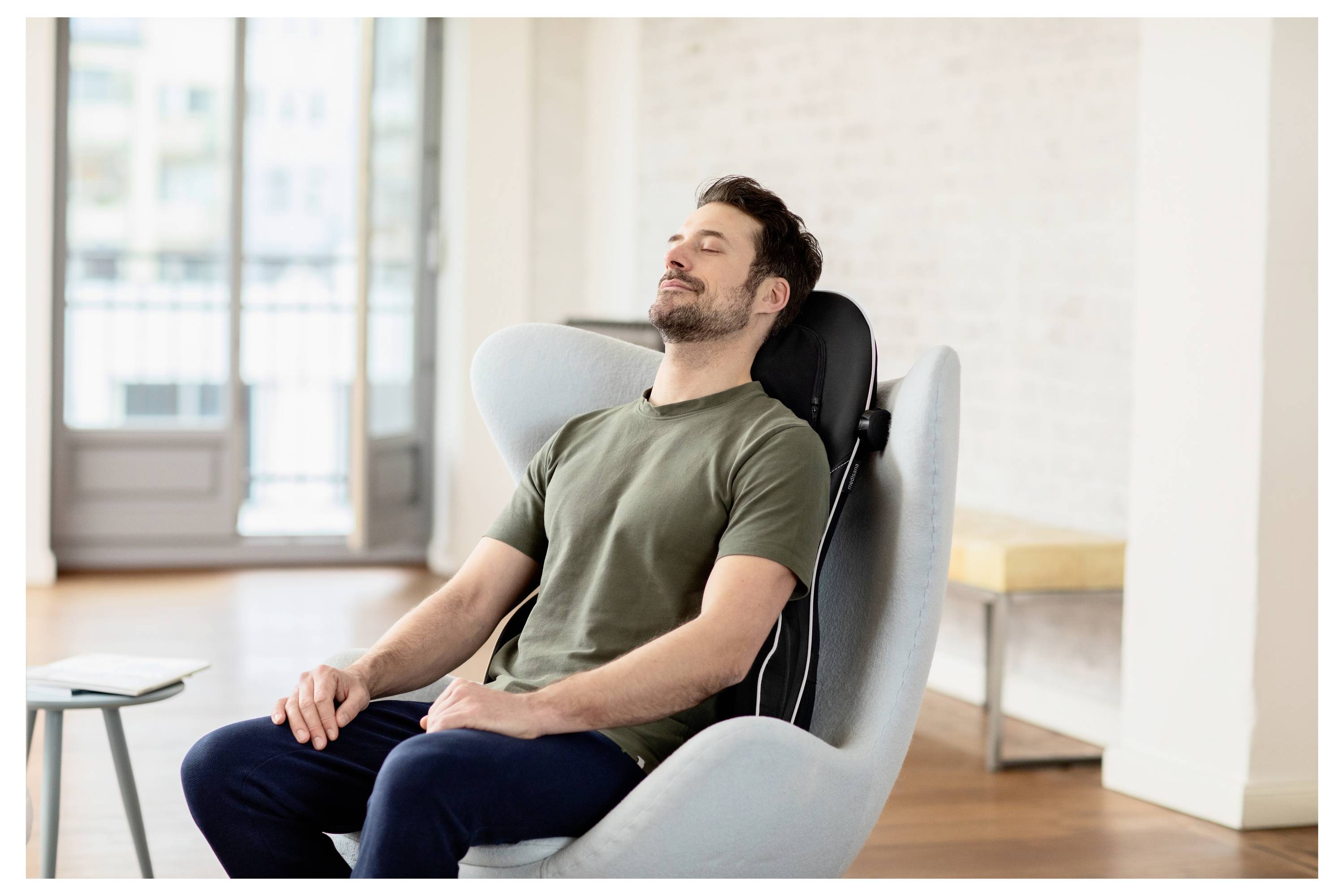 Ein Mann sitzt entspannt in einem modernen Stuhl, die Augen geschlossen, in einem hellen und geräumigen Raum mit Holzböden und großen Fenstern.