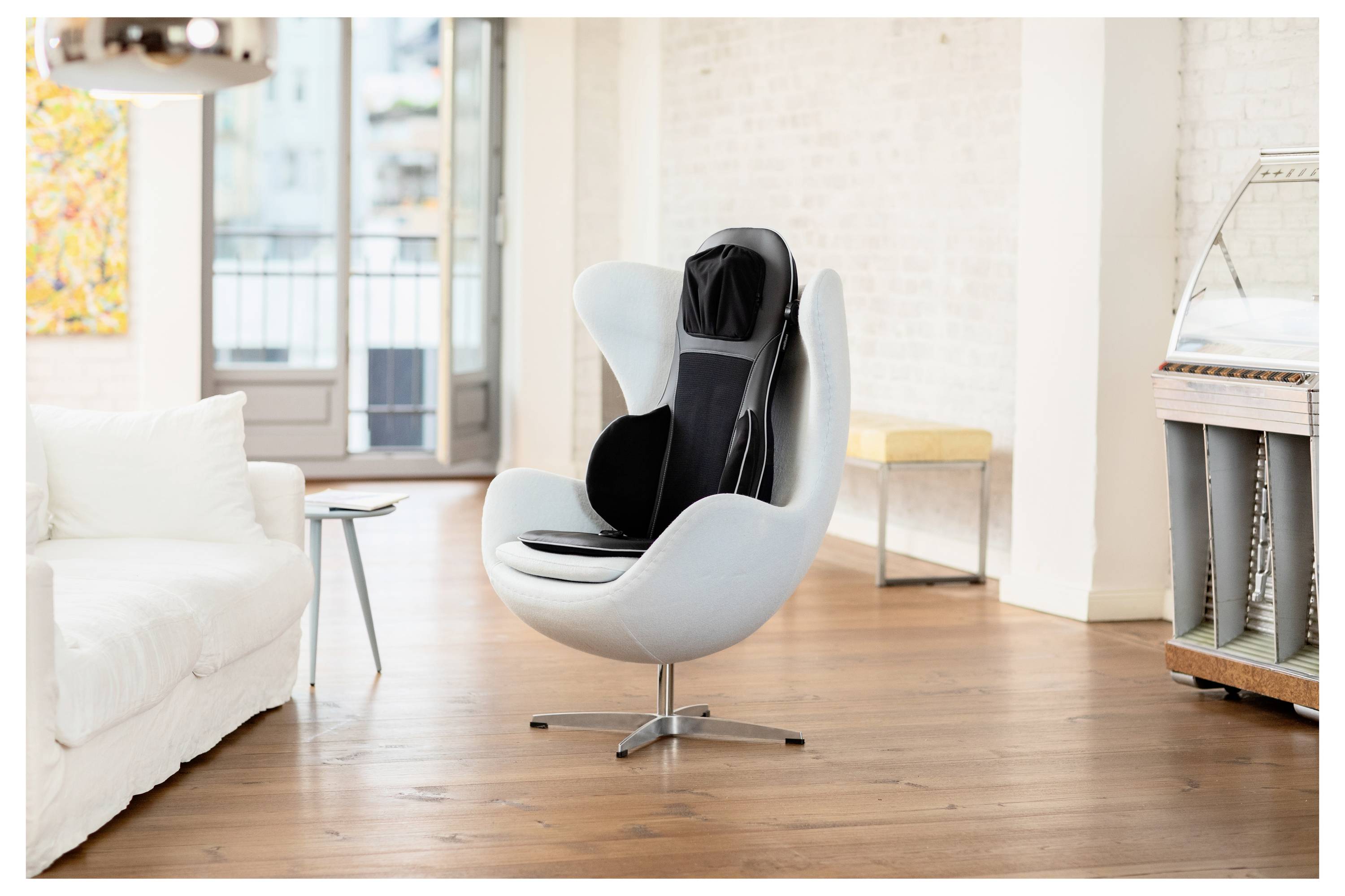 Ein Massagesitzauflage mit elegantem Design wird auf einem modernen, weißen Drehstuhl in einem hellen, zeitgenössischen Wohnzimmer platziert.