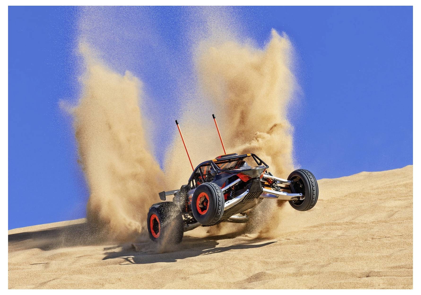 Ein ferngesteuertes Auto mit roten Rädern wirbelt Sand auf, während es einen Wüstenhügel unter einem klaren blauen Himmel erklimmt.