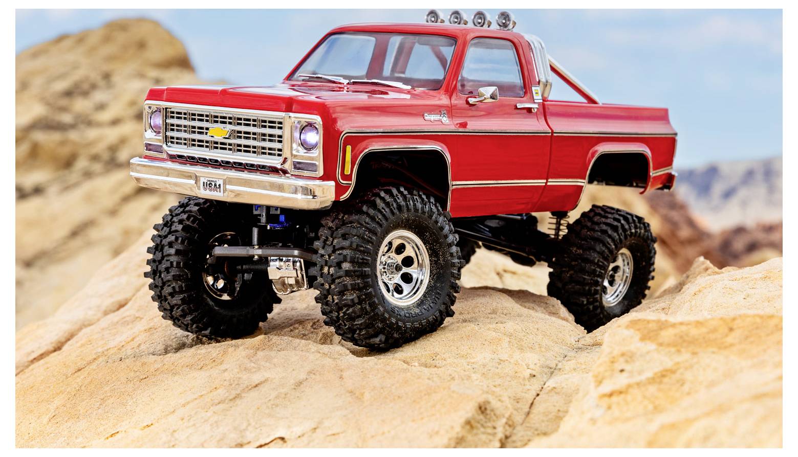 Ein roter Spielzeug-Monstertruck mit großen Reifen ist auf felsigem Gelände positioniert und demonstriert seine Geländegängigkeit. Der Hintergrund zeigt eine Wüstenlandschaft.