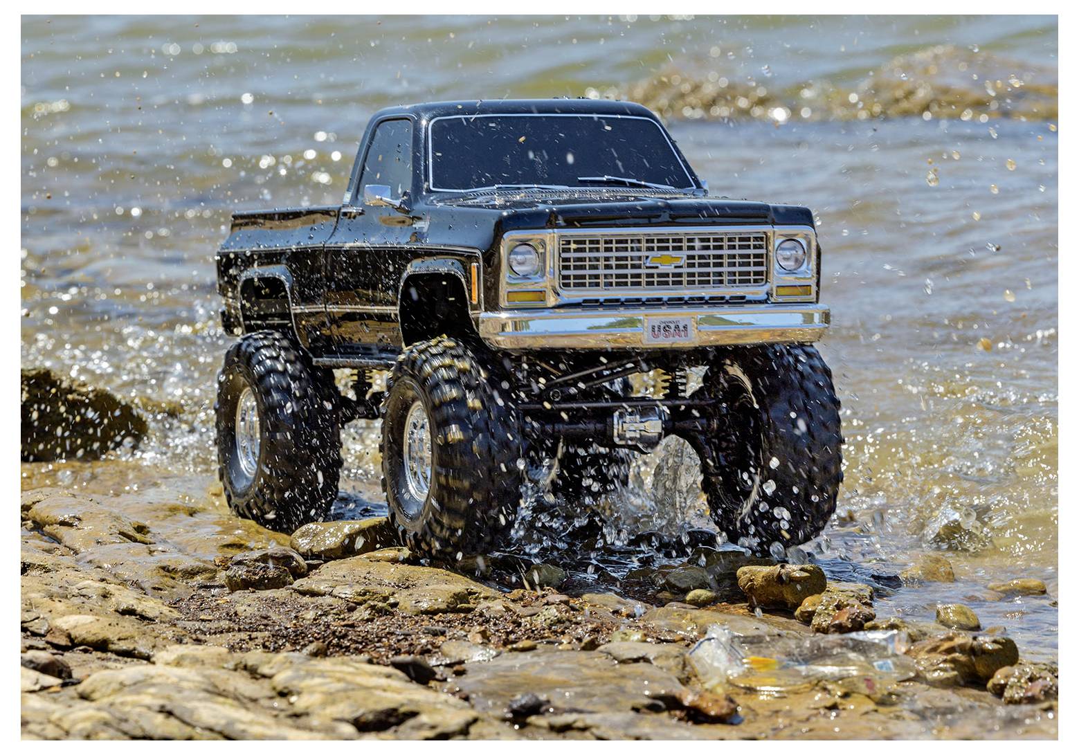 Ein ferngesteuerter Monstertruck fährt über felsiges Gelände an einem See entlang und spritzt Wasser um seine großen Reifen herum.