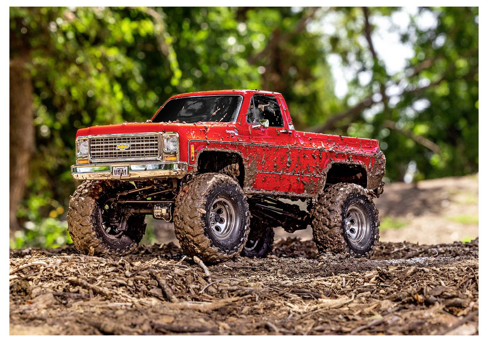 Ein roter Geländewagen-RC-Truck ist mit Schlamm bedeckt und parkt auf einem Schotterpfad, umgeben von Grünpflanzen, was sein robustes Aussehen und seine abenteuerliche Nutzung unterstreicht.