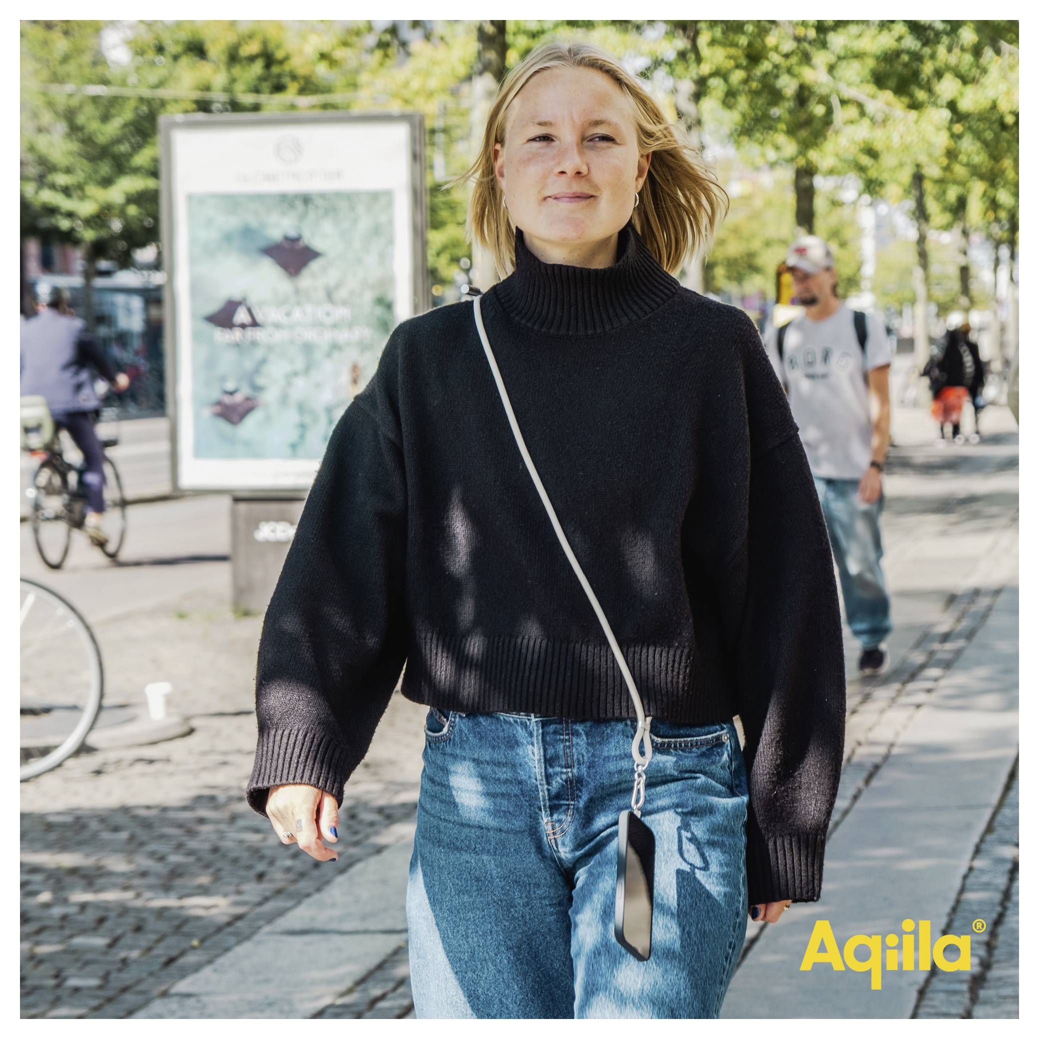 Eine Person, die an einem sonnigen Tag auf einer Straße geht, trägt einen schwarzen Pullover und Jeans und hält eine Tasche. Werbeplakate und Menschen sind im Hintergrund.