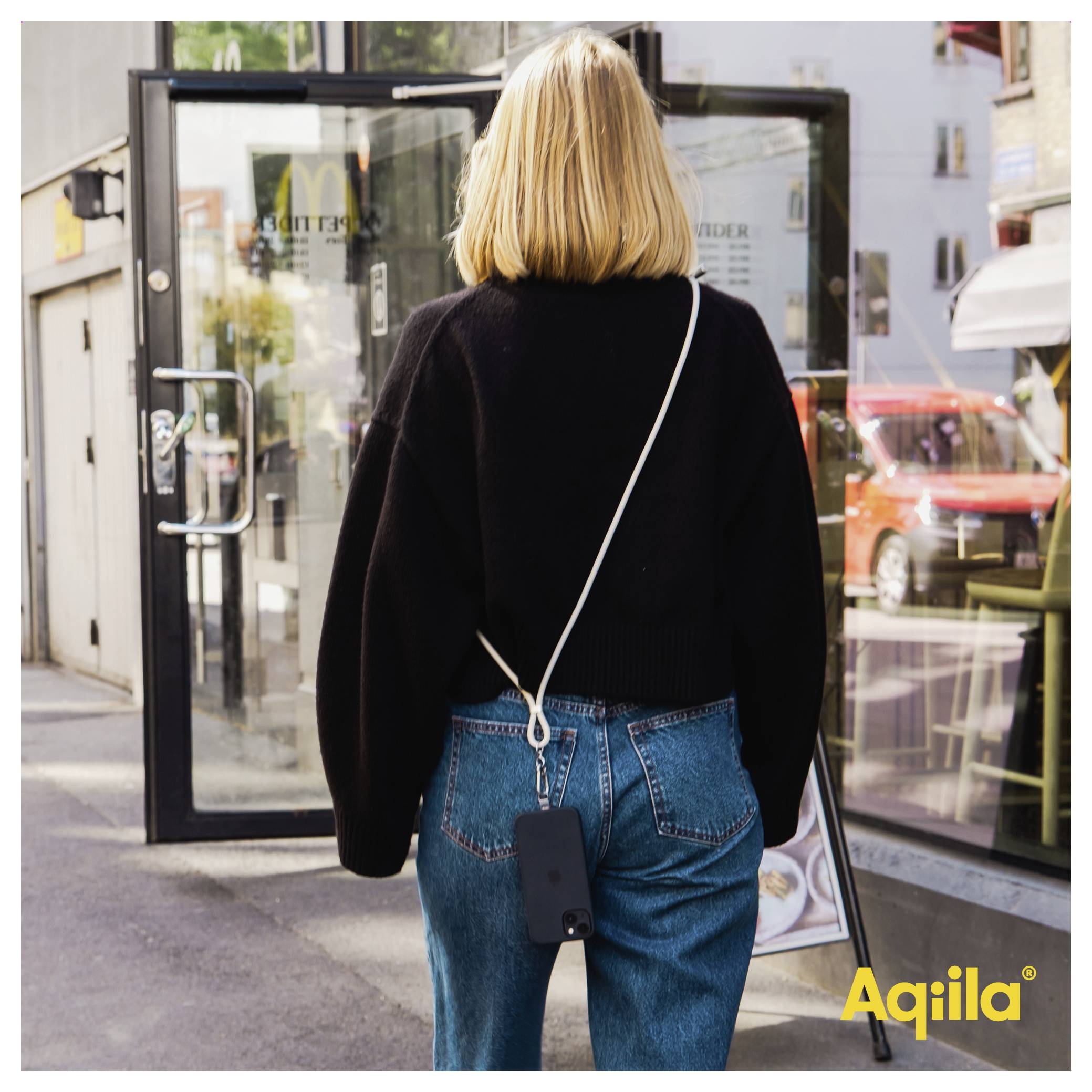 Eine Person mit blonden Haaren, die einen schwarzen Pullover und Jeans trägt, geht in einer urbanen Umgebung auf eine Glastür zu und hält dabei ein Telefon.