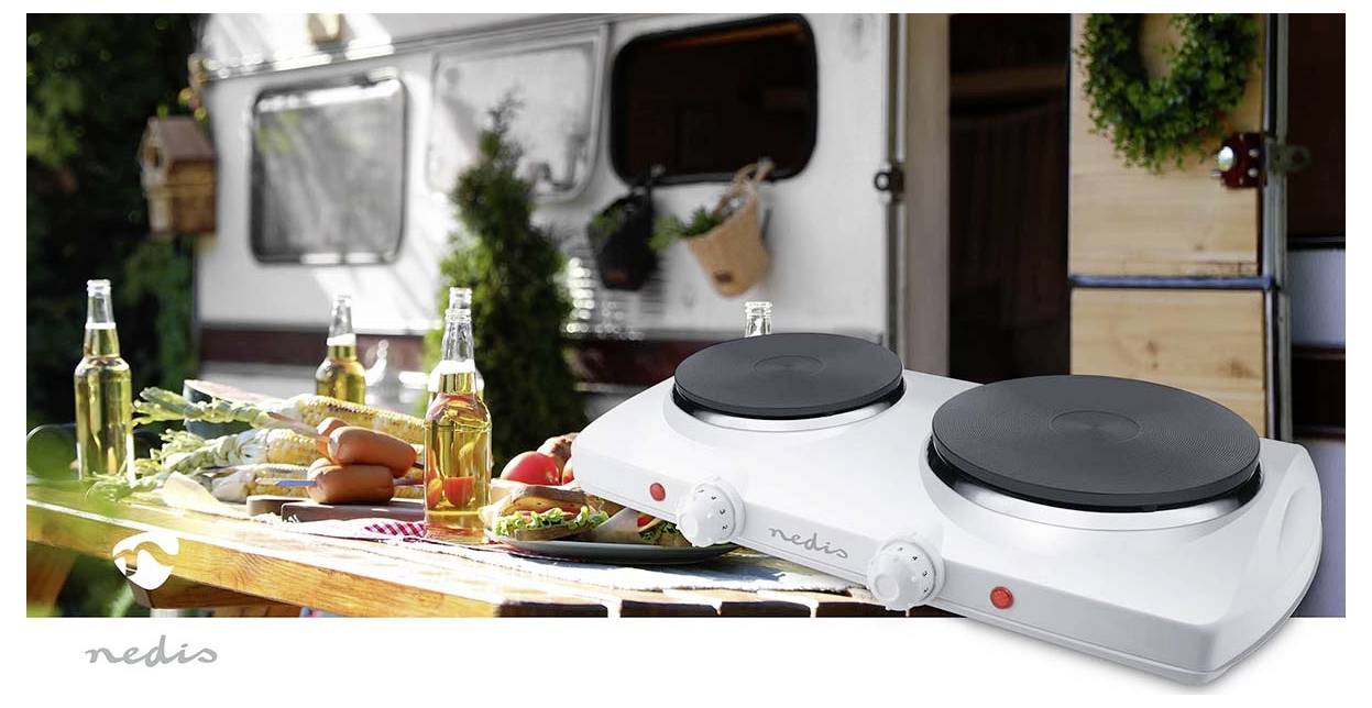 Zweibrenner-Elektroherd auf einem Tisch mit Outdoor-Hintergrund. Flaschen und Lebensmittel befinden sich in der Nähe. Camper und Grünflächen sind zu sehen.