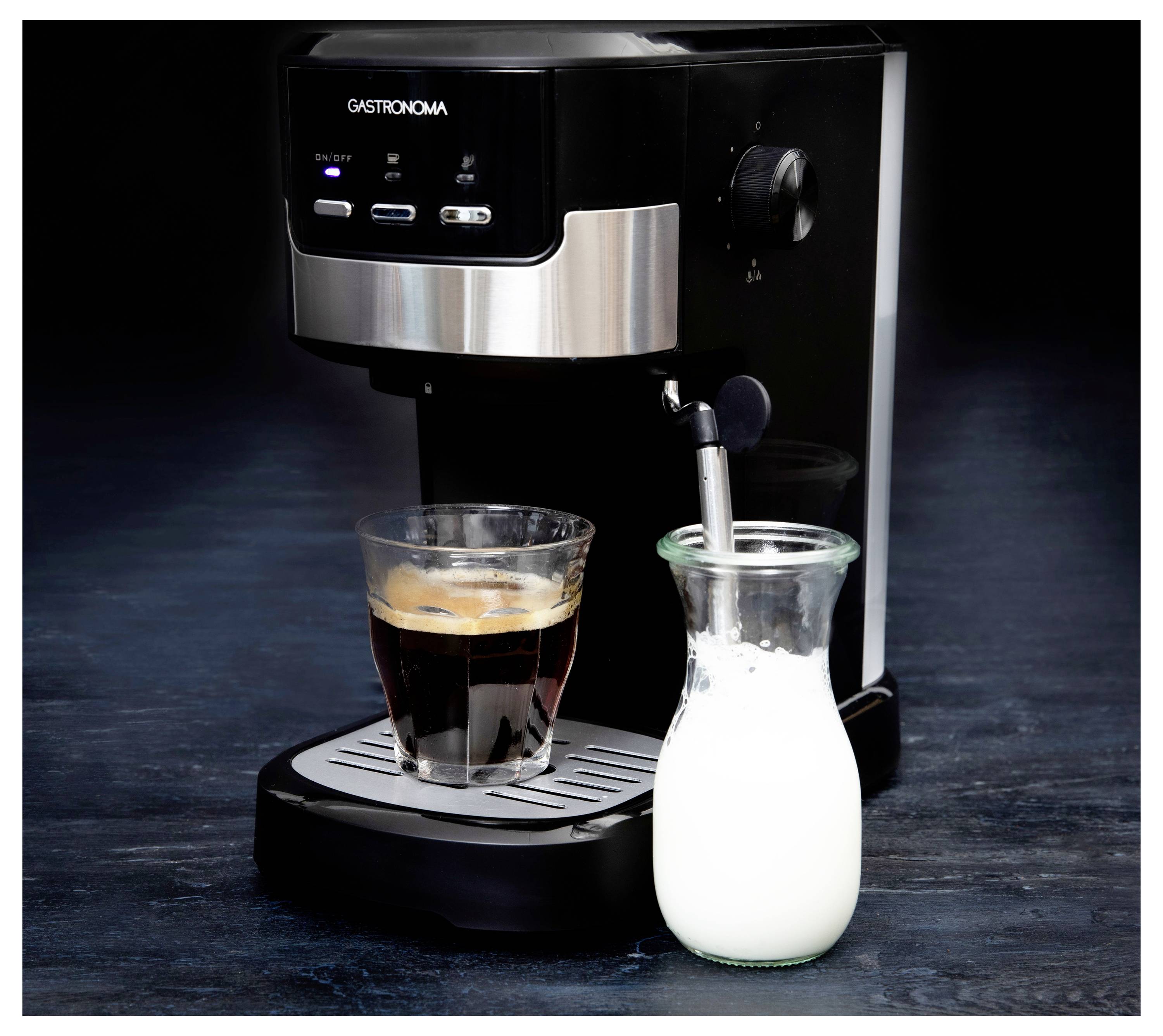 Eine Espressomaschine mit einem Glas Kaffee auf dem Tablett und einem Glaskanne für Milchschaum daneben. Die Maschine ist schwarz und silber.