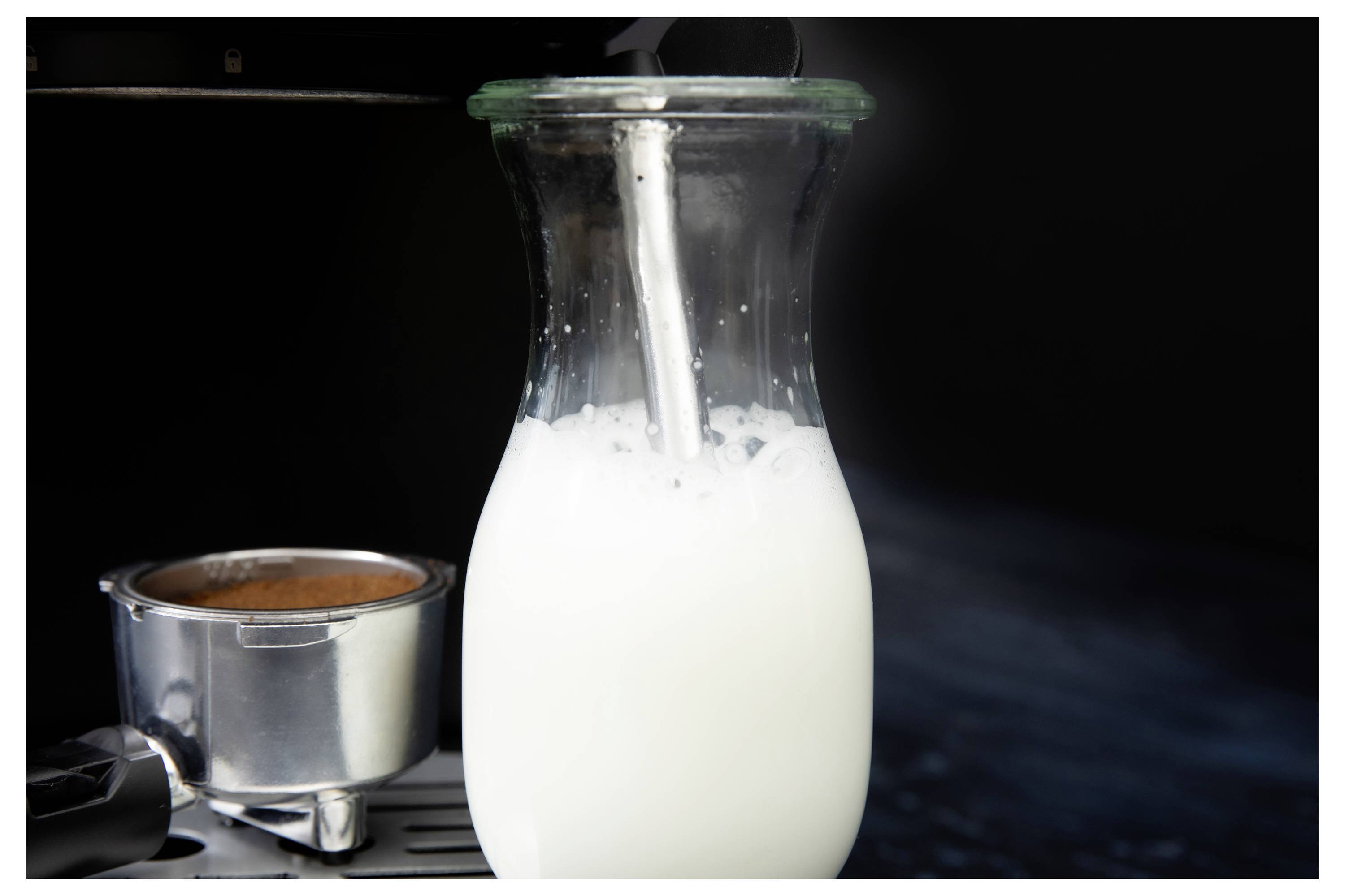 Eine Glaskaraffe, gefüllt mit Milch, wird auf einer Espressomaschine gedämpft, neben einem Siebträger mit Kaffee, vor einem dunklen Hintergrund.