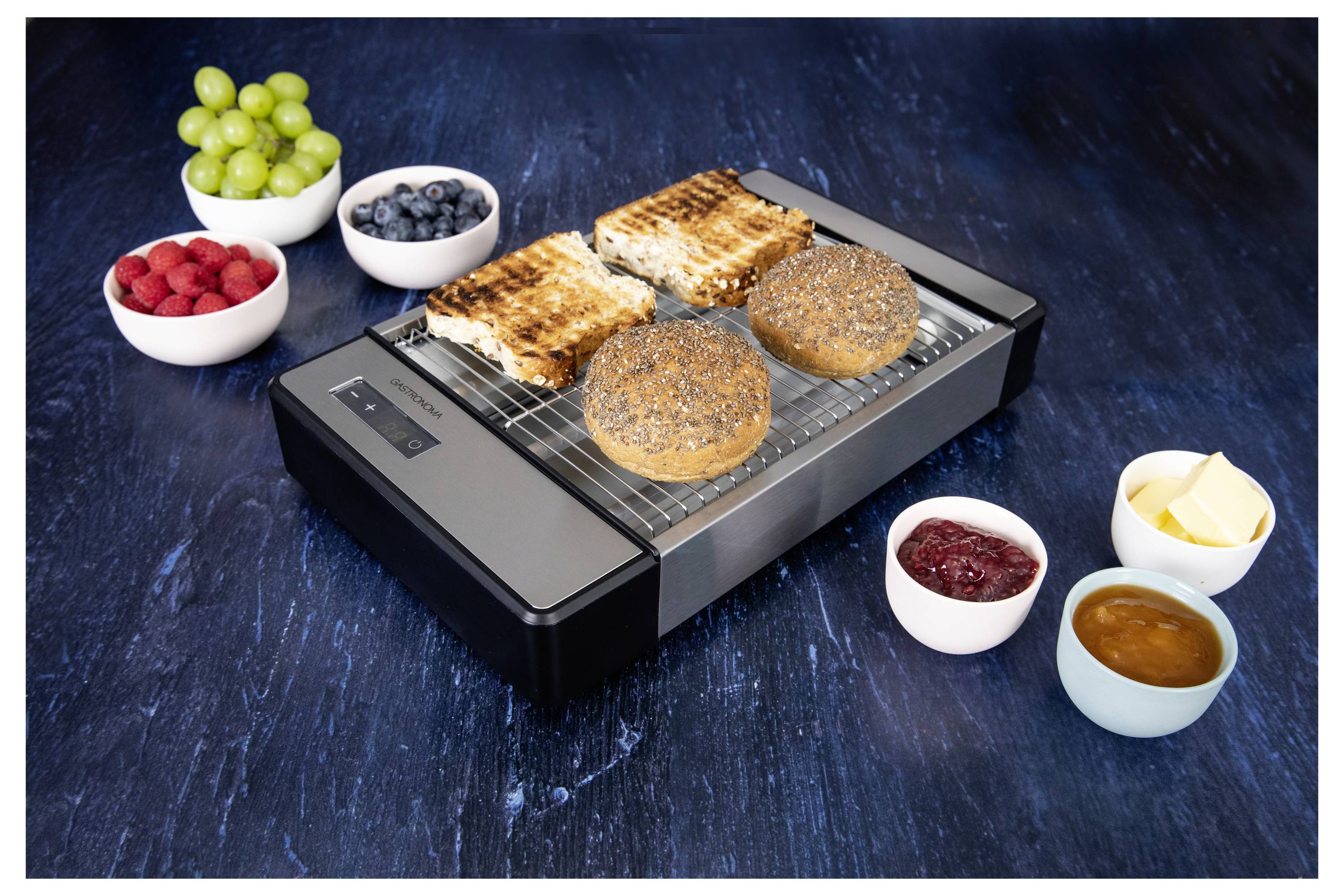 Ein schlanker Wärmebehälter hält getoastete Sandwiches und Bagels. Umgeben wird er von Schalen mit Trauben, Blaubeeren, Himbeeren, Butter, Marmelade und Sauce.