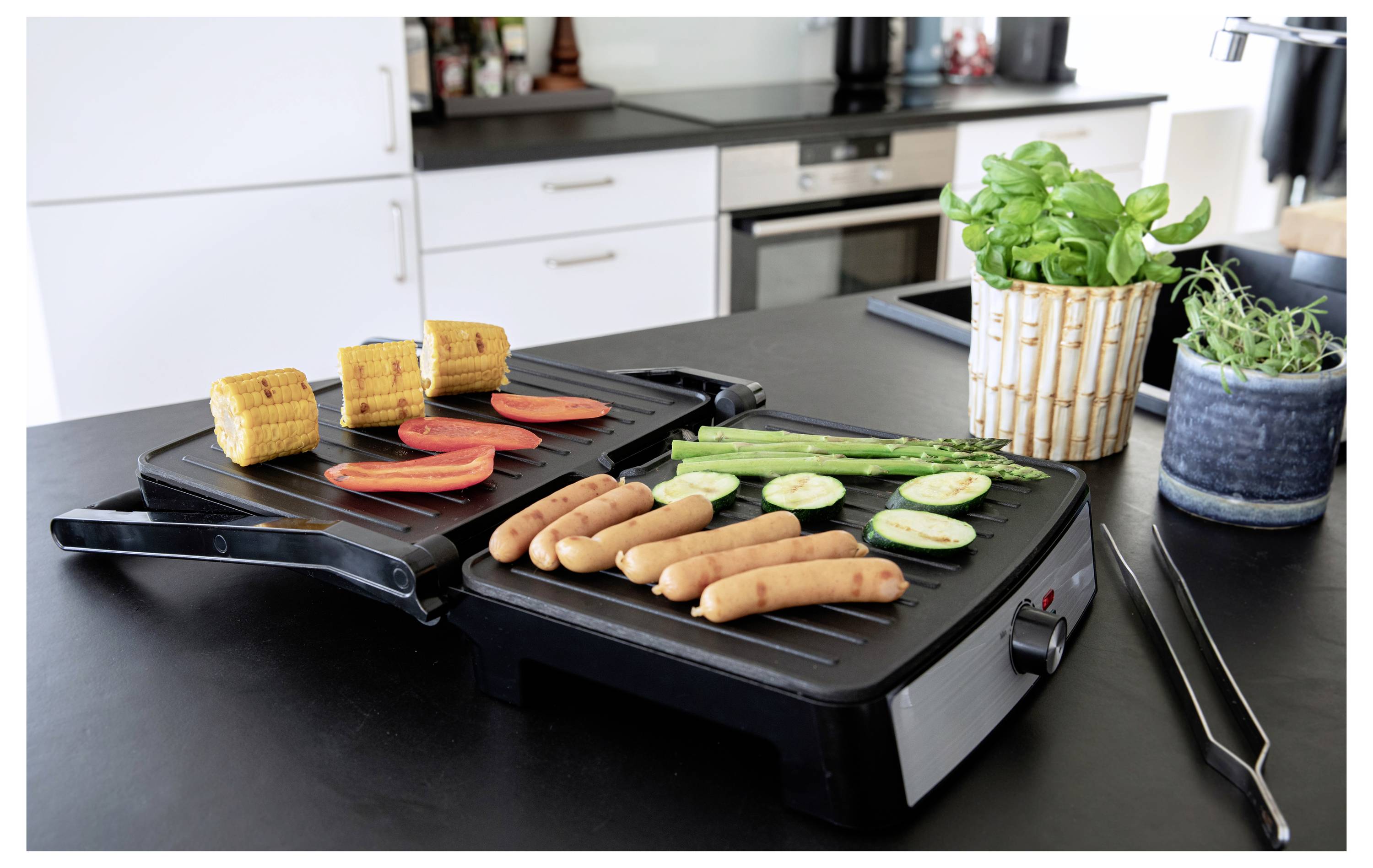 Ein offener Grill auf einer Küchenarbeitsplatte mit Mais, Tomaten, Würstchen, Zucchini und Spargel. In der Nähe stehen Kräutertöpfe und eine moderne Kücheneinrichtung.
