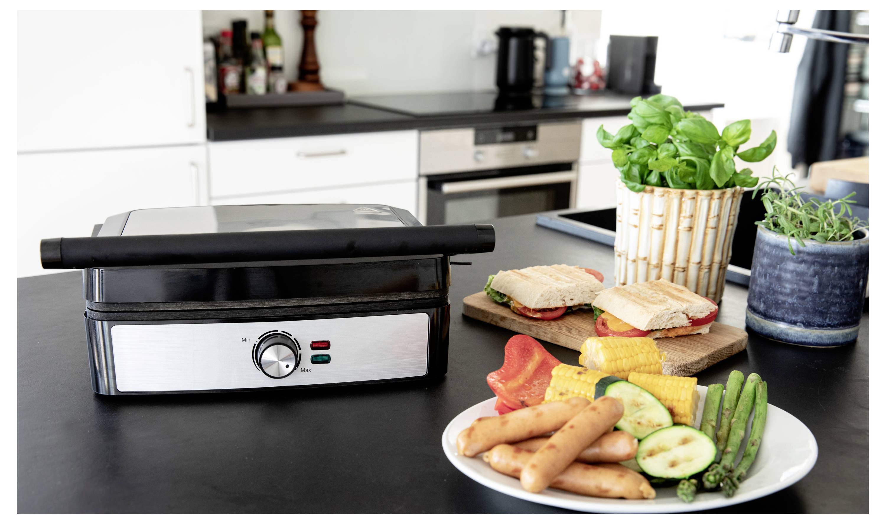 Grill auf der Arbeitsplatte in einer Küche mit zwei Paninis, Pflanzen und einem Teller mit Gemüse, darunter Paprika, Mais, Spargel und Zucchini.