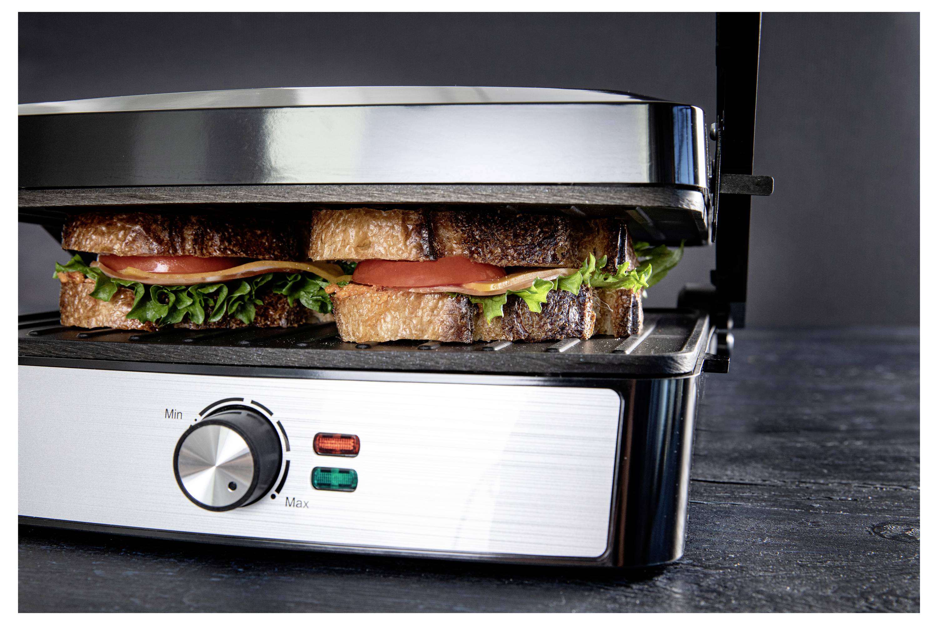 Ein Panini-Grill, der zwei Sandwiches mit Salat, Tomate und Käse grillt, steht auf einer dunklen Arbeitsplatte. Die Bedienknöpfe zeigen Einstellungen von minimal bis maximal.
